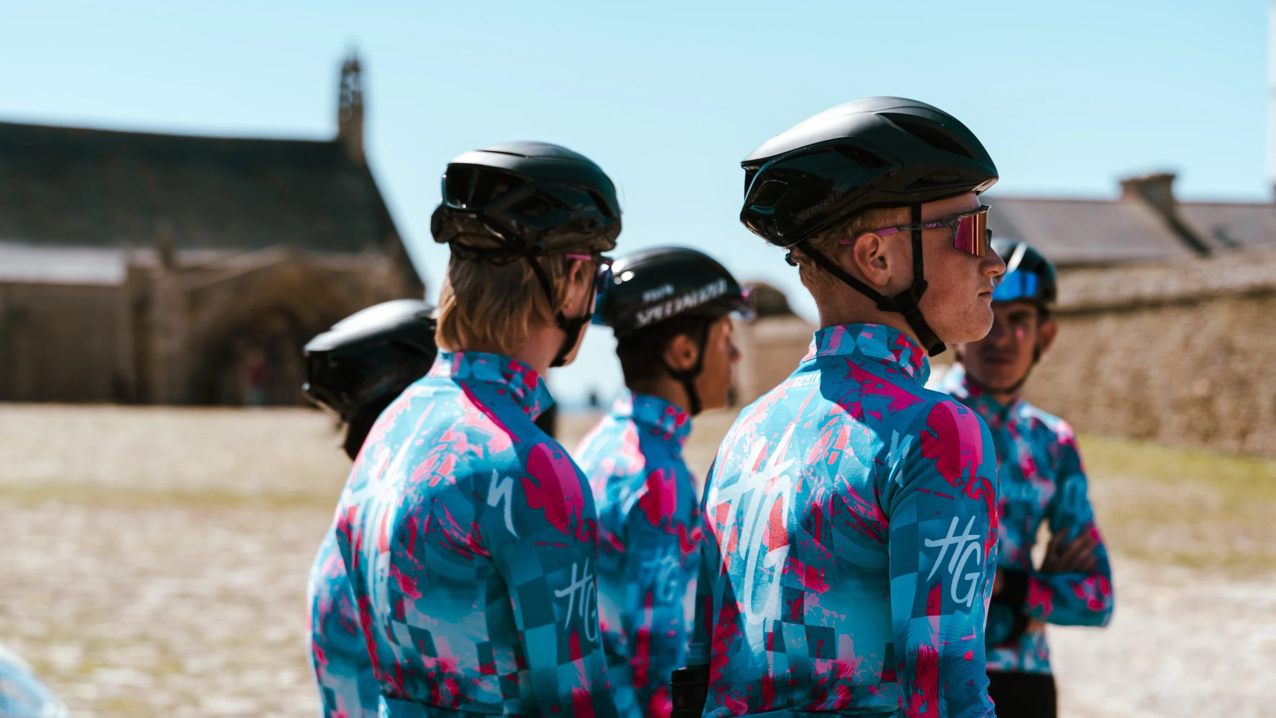 Groupe de personnes portant des casques noirs, habillées d'un maillot coloré à motifs bleus et roses, probablement des cyclistes, en ligne lors d'une activité en plein air sous un ciel clair.
