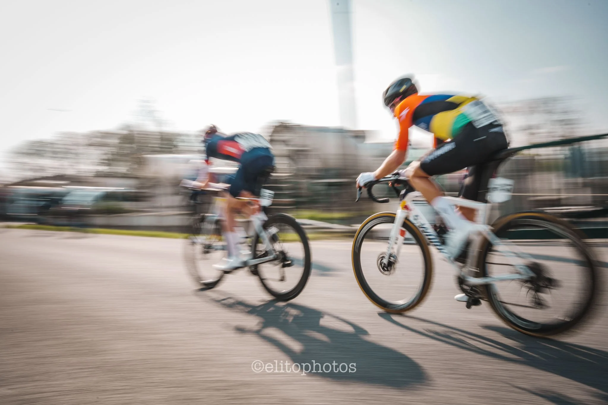 Deux cyclistes en mouvement rapide sur une route en milieu urbain, flous en raison de la vitesse, sous un ciel clair.