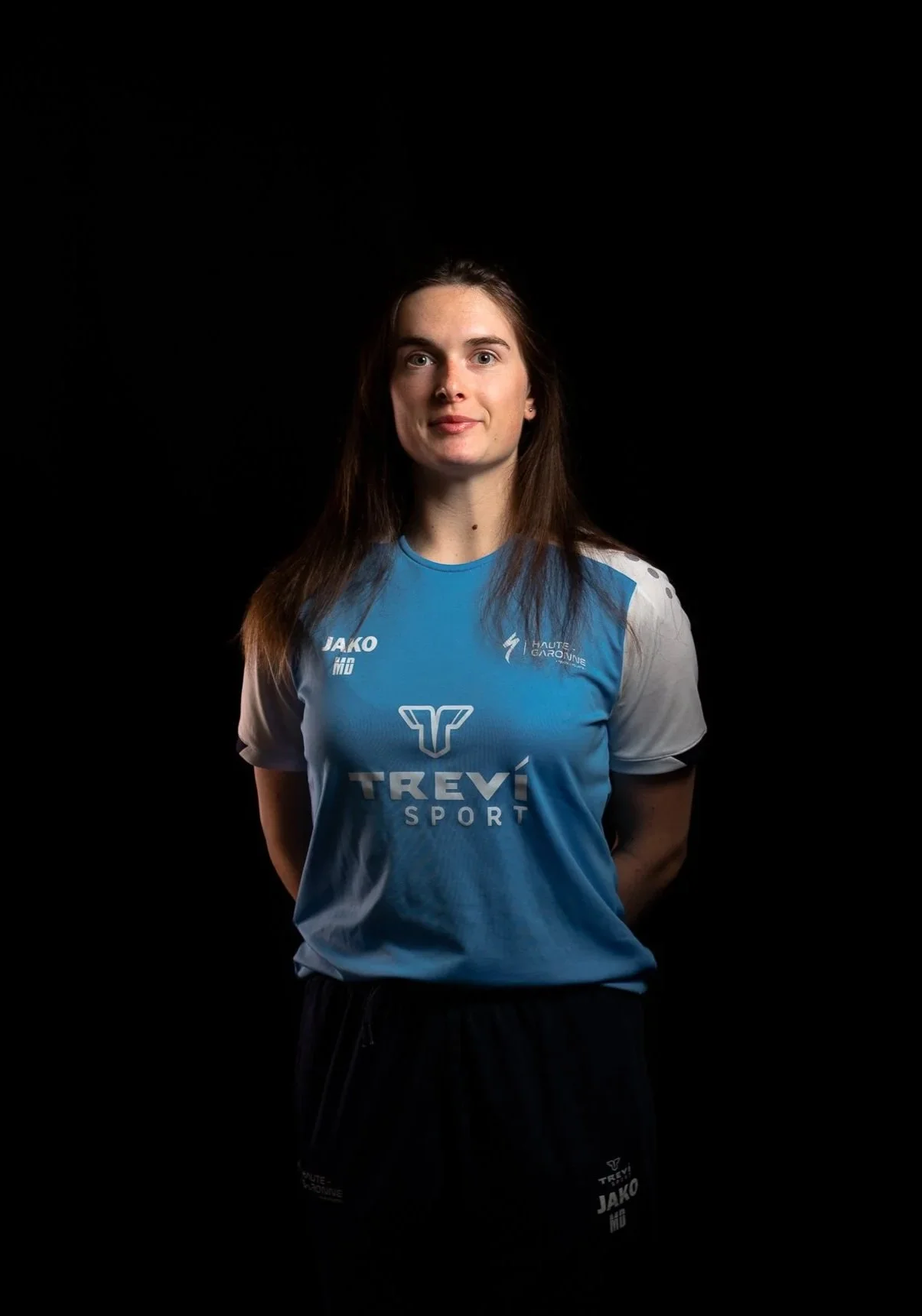 Jeune femme souriante en tenue de sport bleue et blanche, debout, fond noir, portant un maillot avec le logo Trevi Sport, dans une séance photo studio.