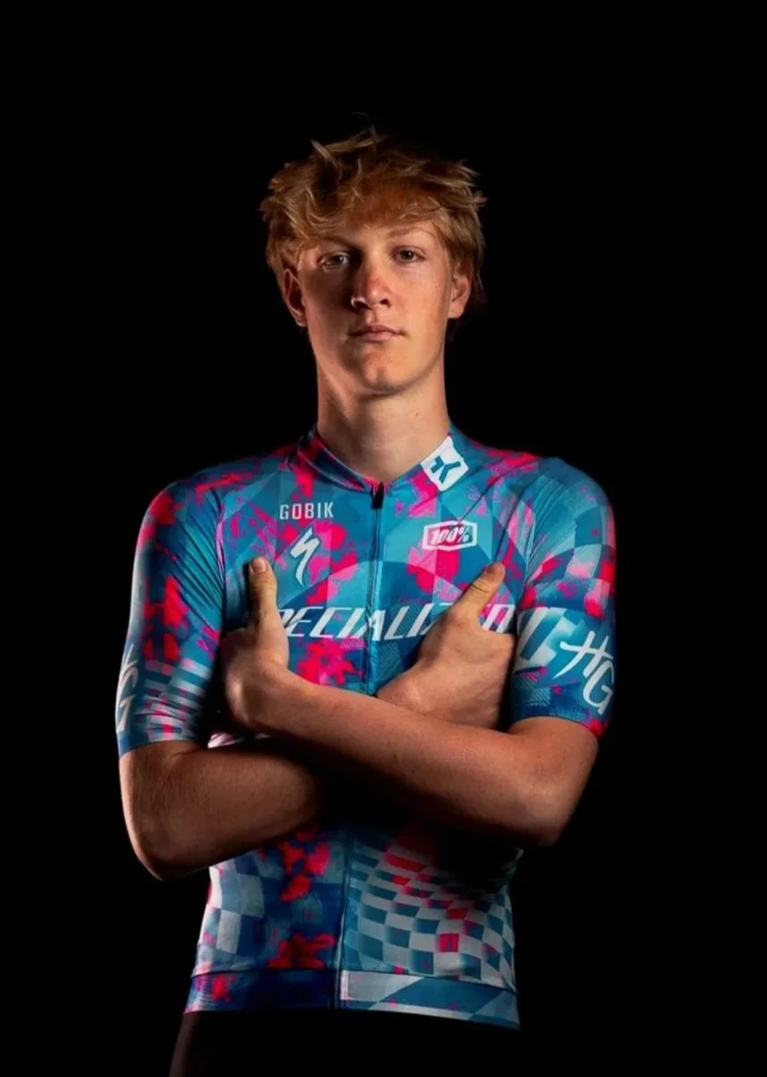 Un jeune homme en maillot de cycliste coloré, posant avec les bras croisés contre un fond noir.