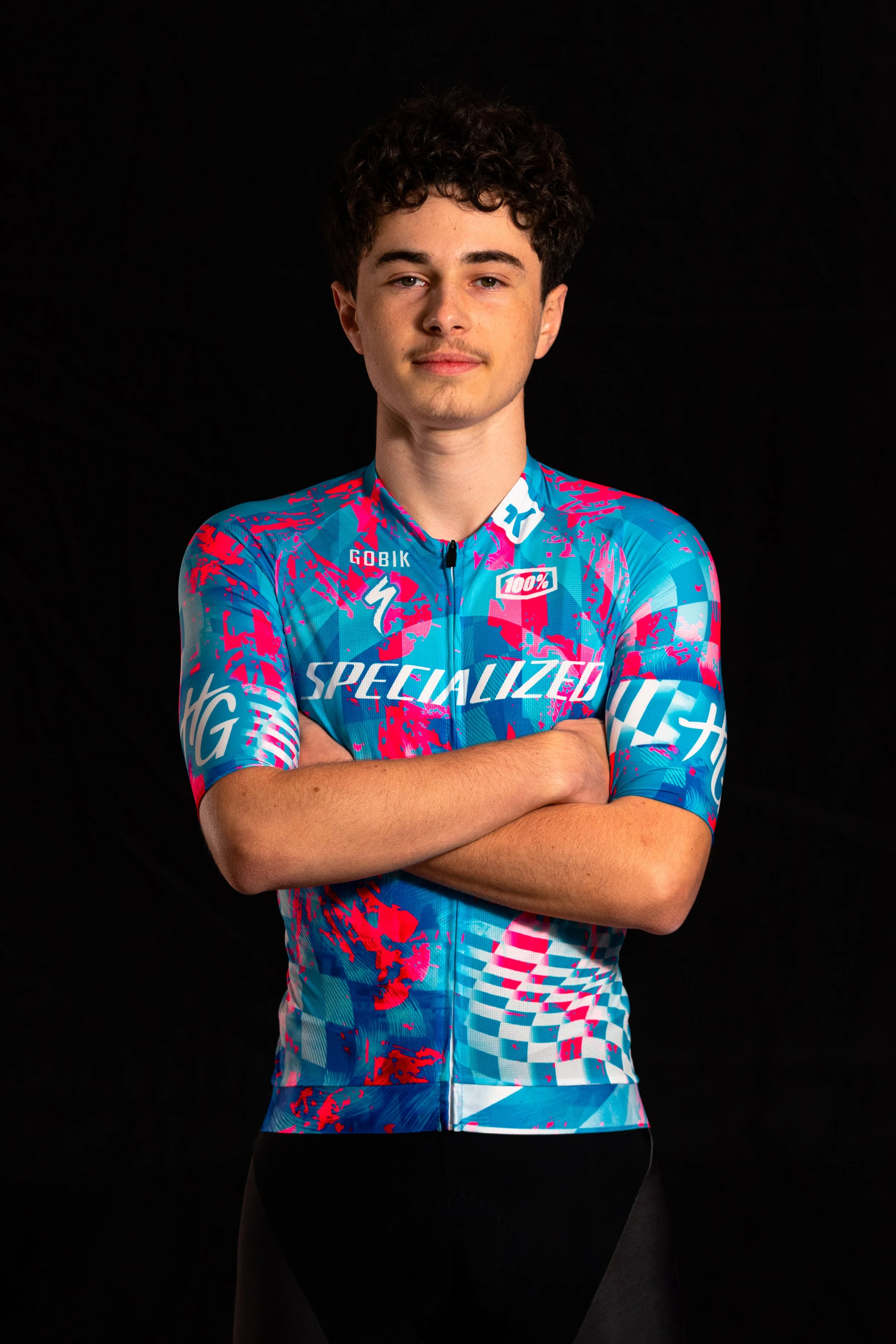 Jeune homme portant un maillot de cyclisme bleu avec des motifs roses et blancs, bras croisés, fond noir.