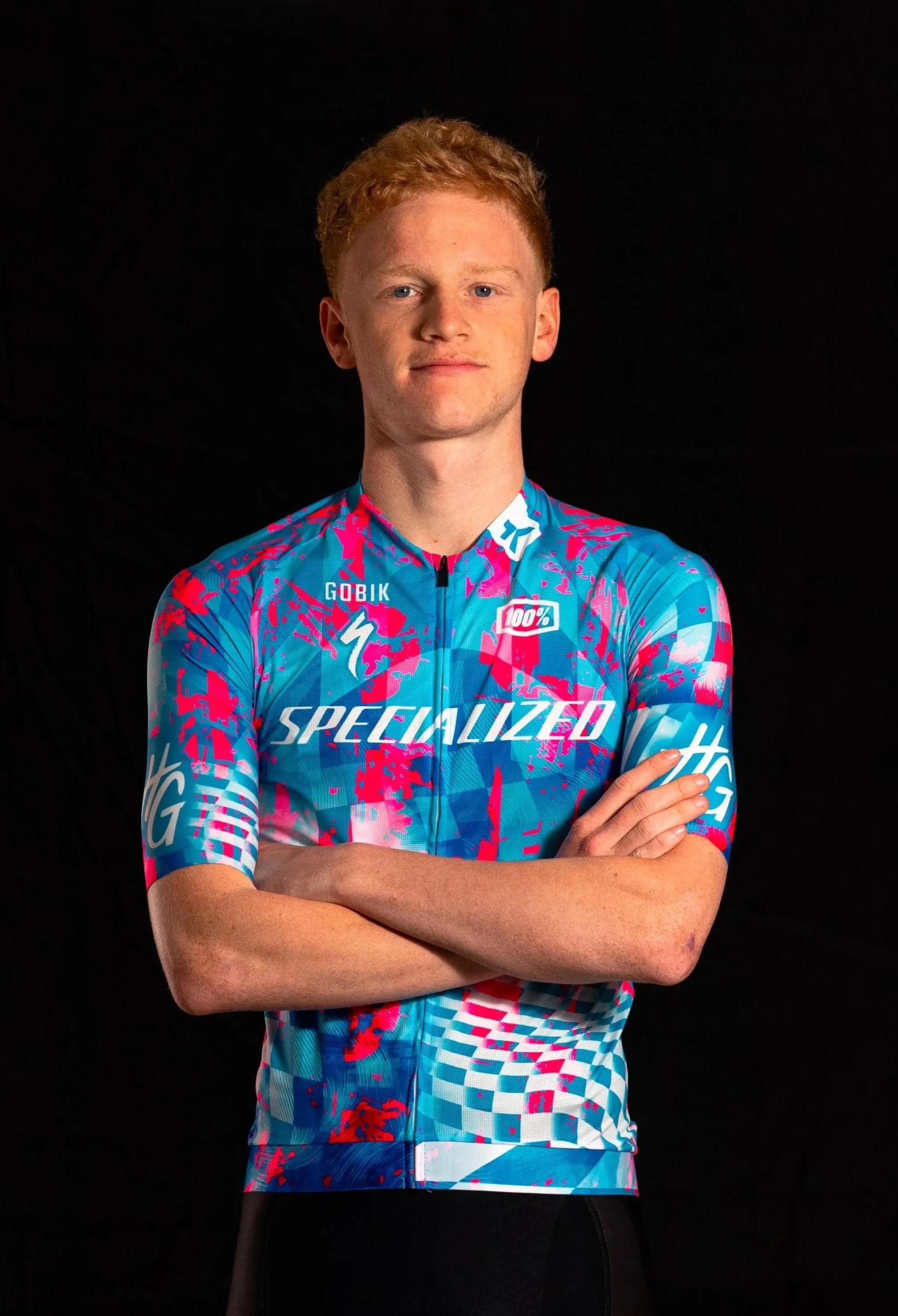 Jeune homme portant un maillot de cyclisme coloré, bras croisés, contre un fond noir.
