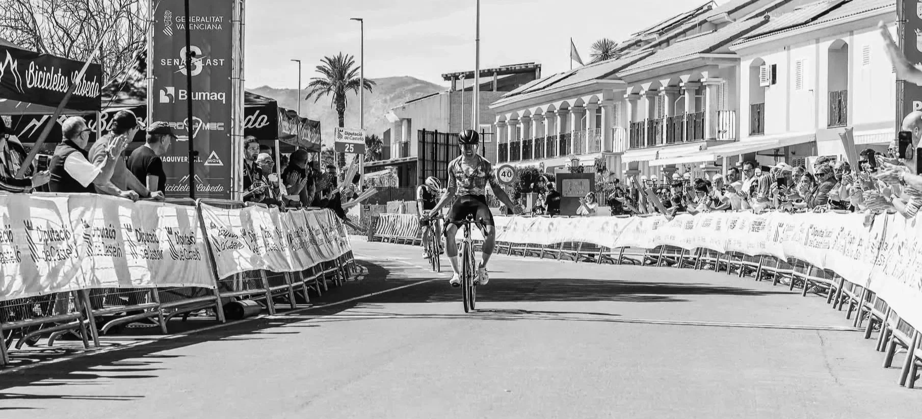 Un coureur à vélo franchit la ligne d'arrivée lors d'une compétition, entouré de spectateurs et de panneaux publicitaires dans une rue urbaine avec bâtiments résidentiels et montagnes en arrière-plan.