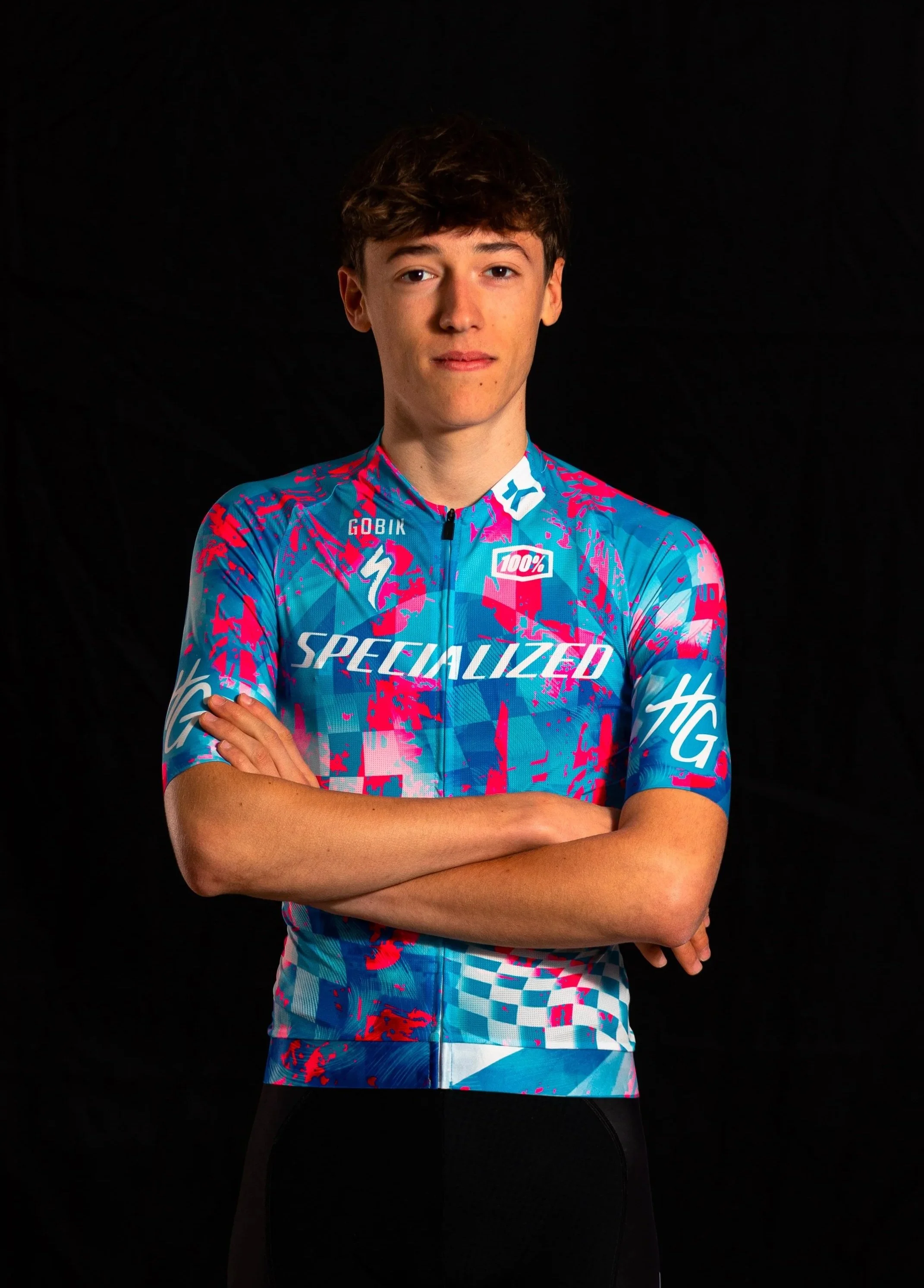 Un jeune homme en maillot de cyclisme coloré, posé contre un fond noir, bras croisés, regardant vers l'appareil.