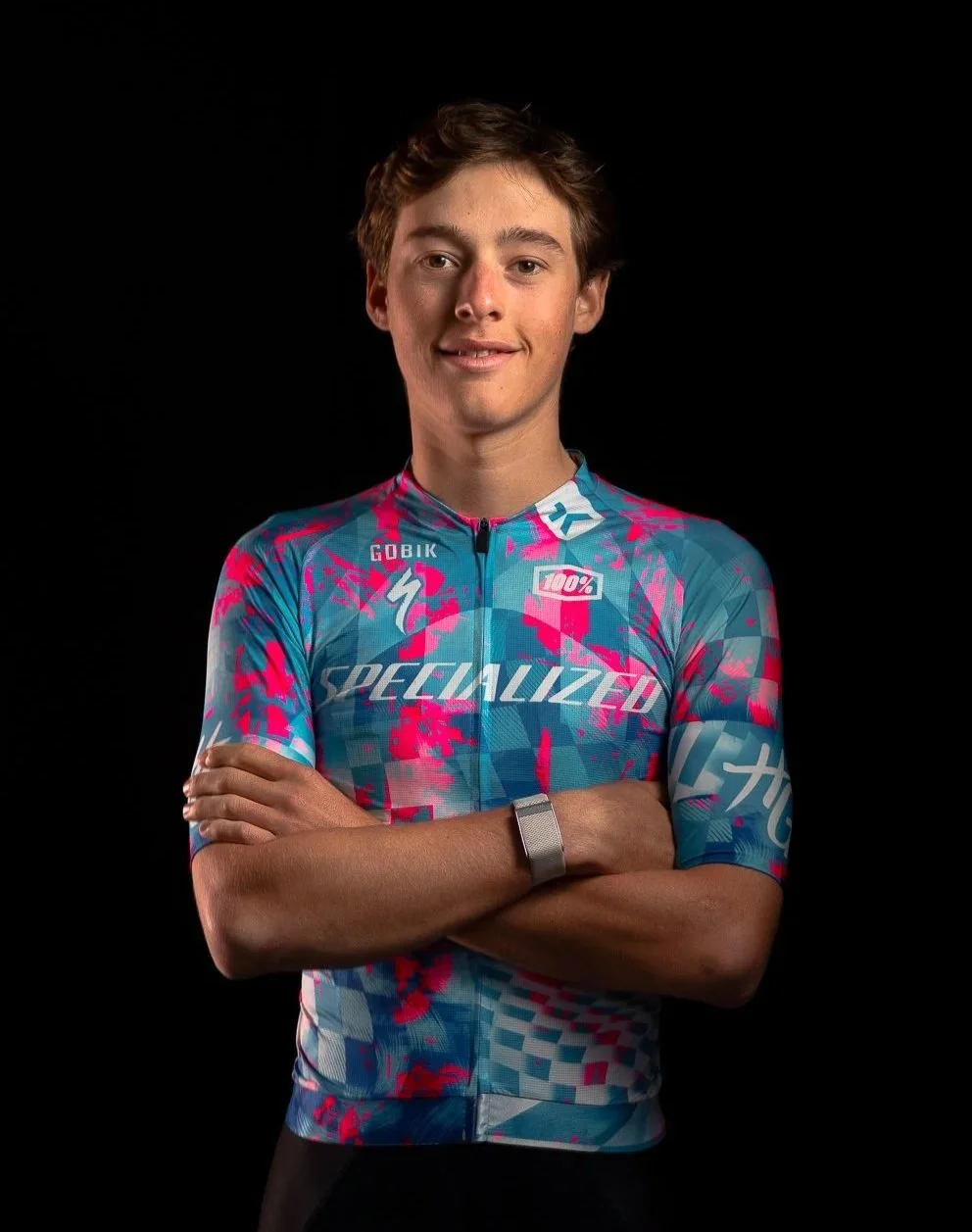 Jeune homme souriant portant un maillot de cyclisme coloré avec le logo 'Specialized' et des motifs roses, bleus et blancs, avec un fond noir.