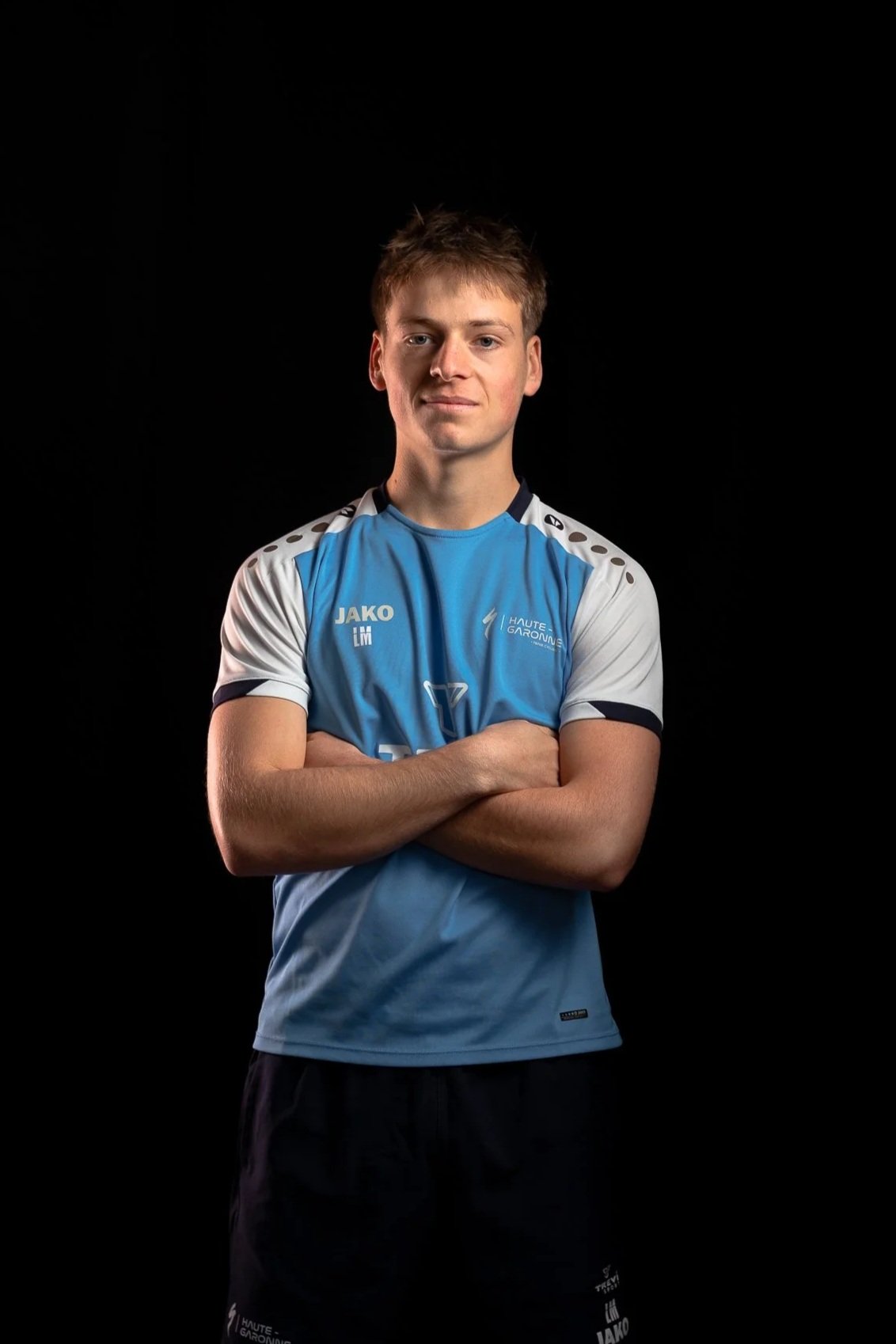 Un jeune homme en tenue de sport bleu clair avec des épaules blanches, se tenant les bras croisés contre un fond noir.