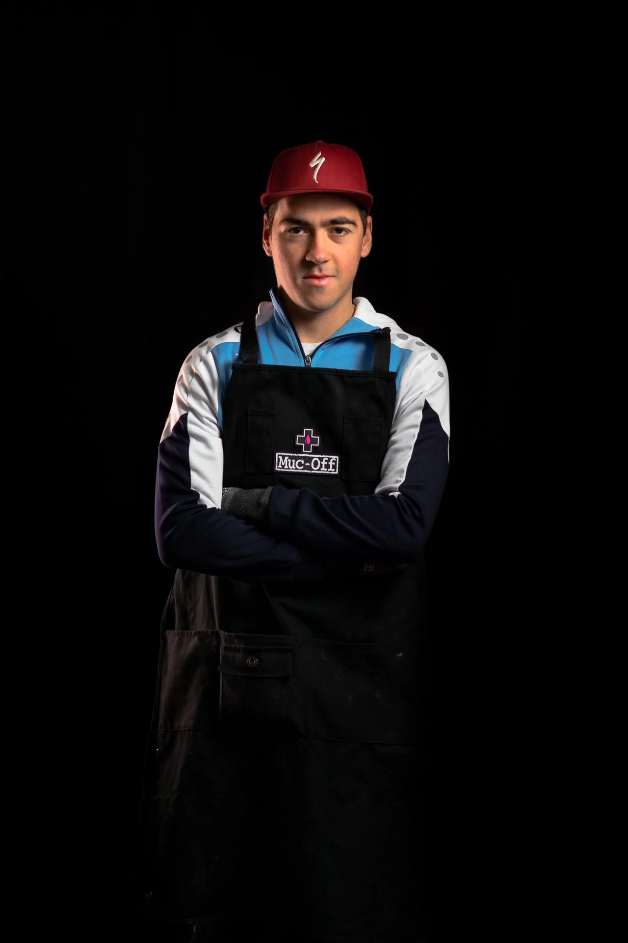 Un jeune homme portant une casquette rouge, un sweat bleu et un tablier noir avec une étiquette "Muc-Off" se tient contre un fond noir.