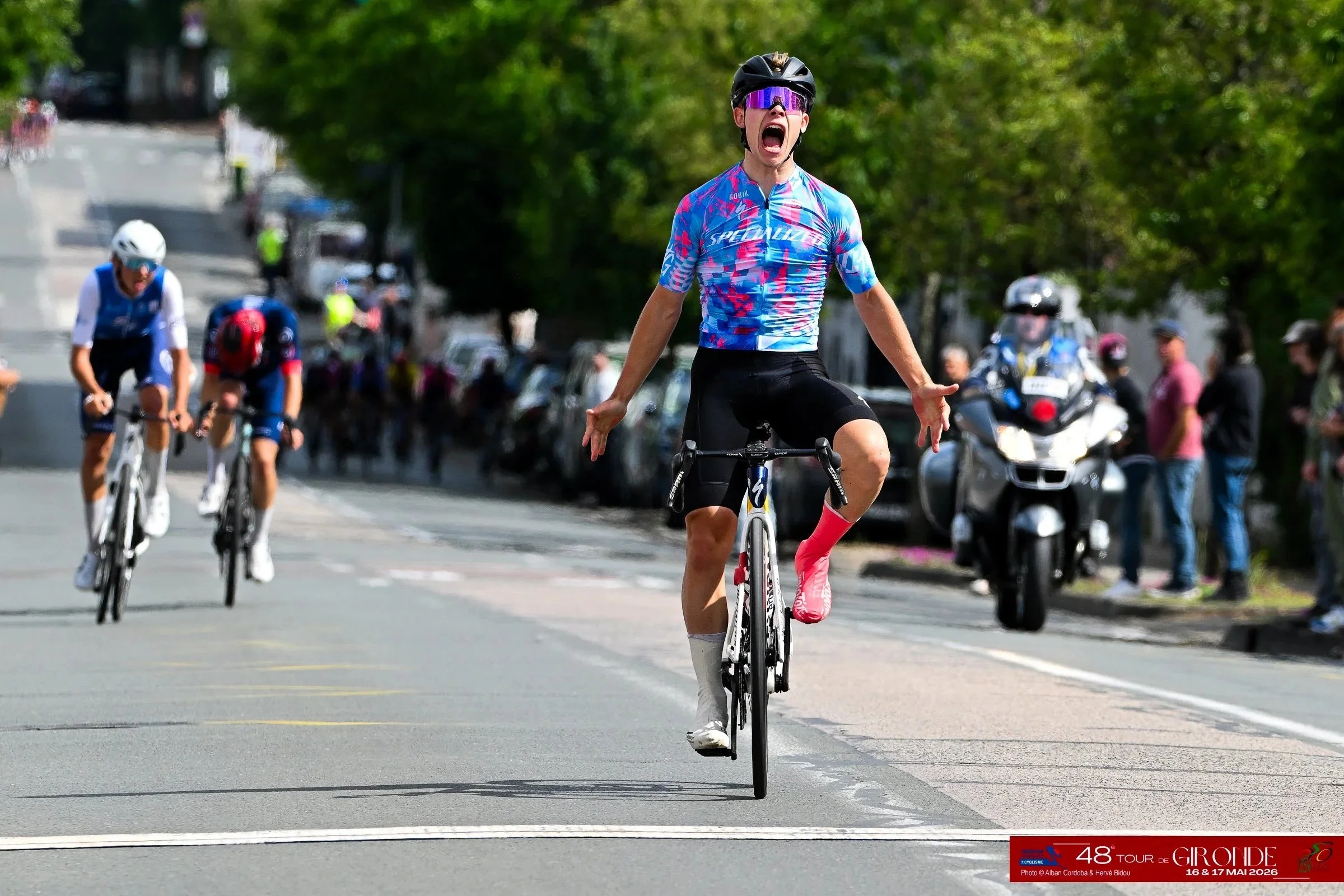 Un cycliste en pleine course, criant de joie ou d'excitation, portant un casque noir, des lunettes de soleil violettes, un maillot de cyclisme bleu avec des motifs roses et bleus, et des chaussettes roses. Derrière lui, d'autres cyclistes et des spectateurs sont visibles, avec des voitures stationnées sur le côté de la route. La scène se déroule le long d'une rue bordée d'arbres sous un ciel ensoleillé.