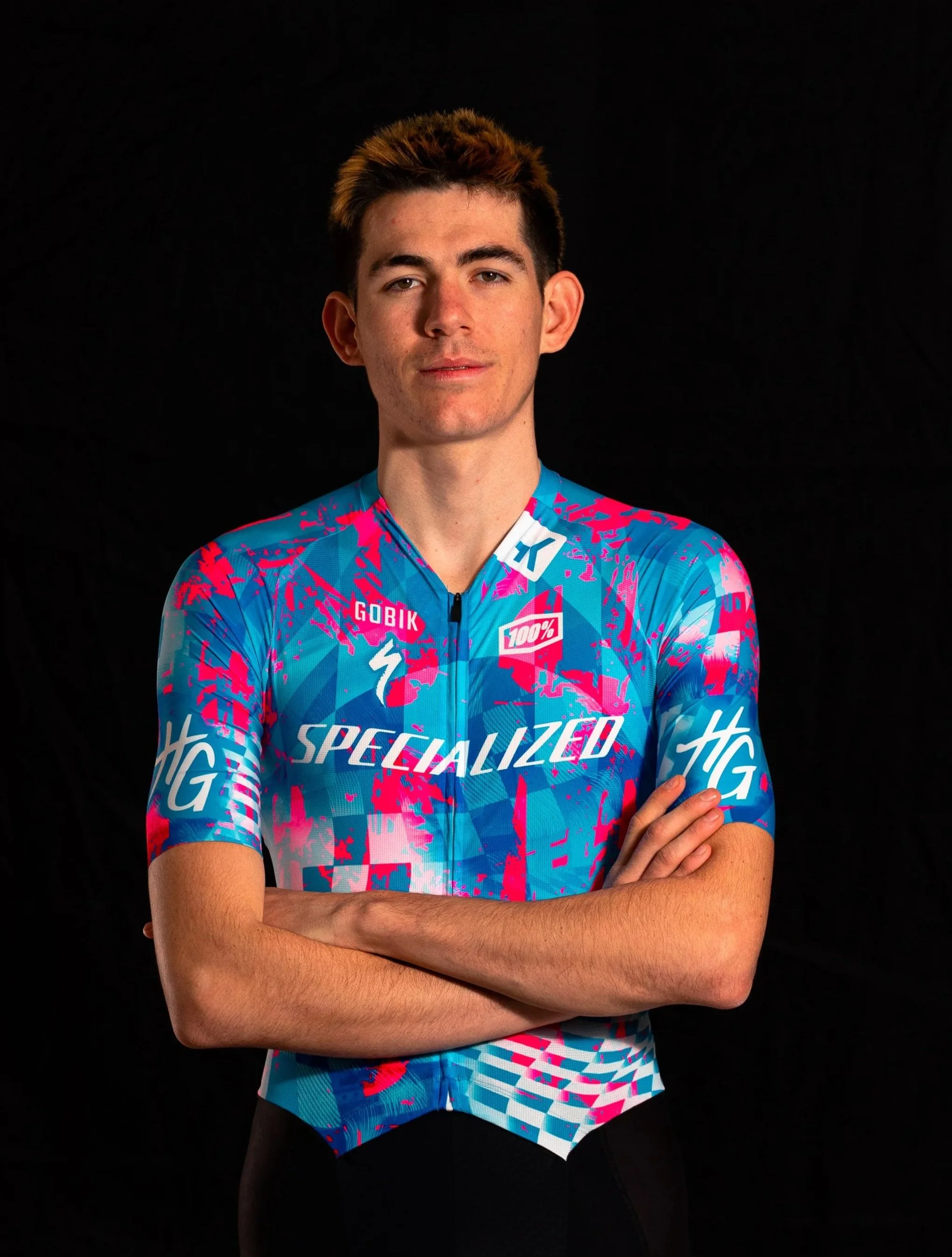 Un jeune homme à cheveux bruns et blonds portant un maillot cycliste coloré avec des logos, bras croisés, contre un fond noir.