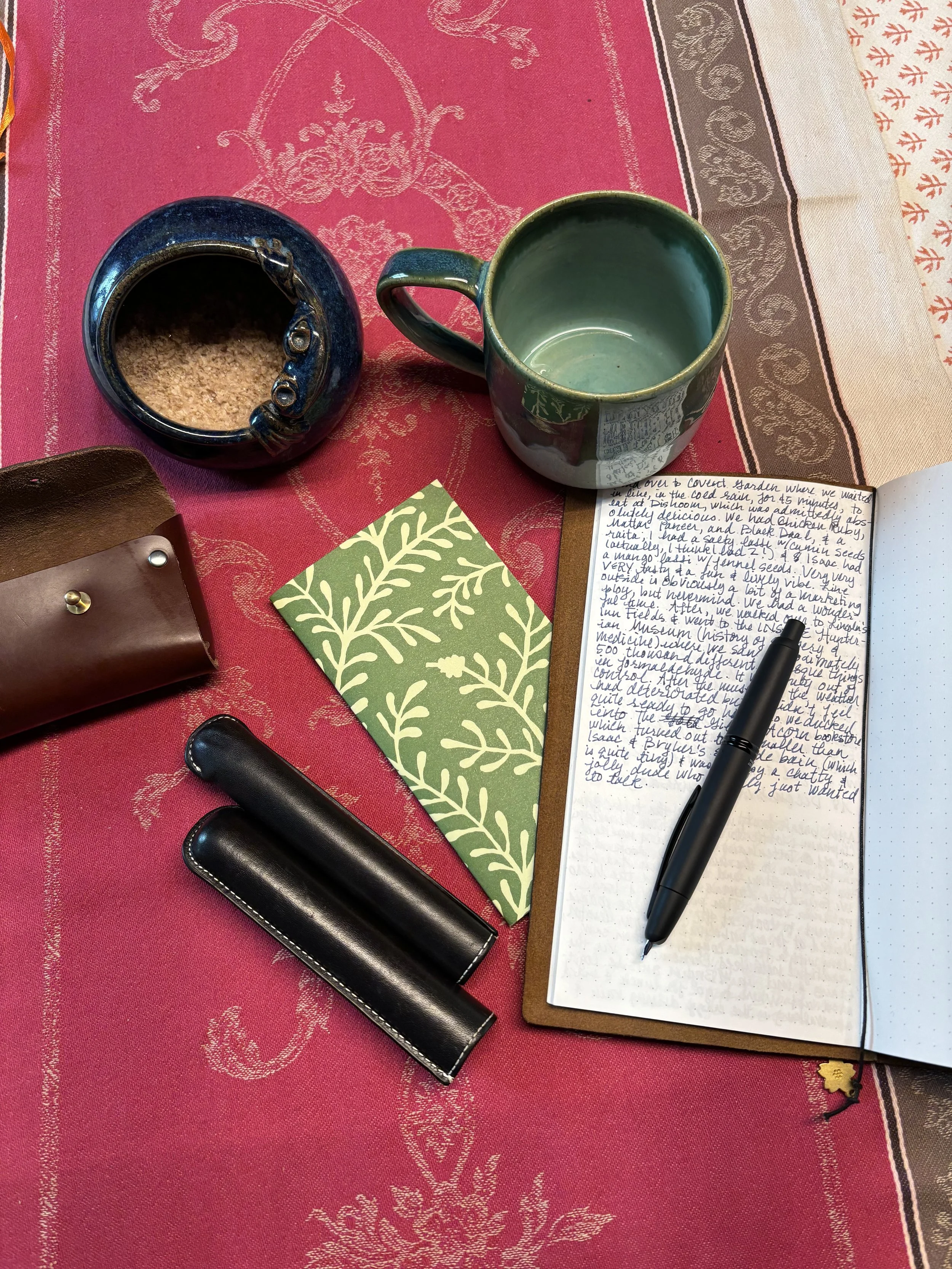 Une table avec une tasse verte vide, un pot avec du sucre, un carnet avec des notes en cursif, un stylo, un portefeuille en cuir et deux étuis à lunettes noirs sur un tissu rouge à motifs anciens.