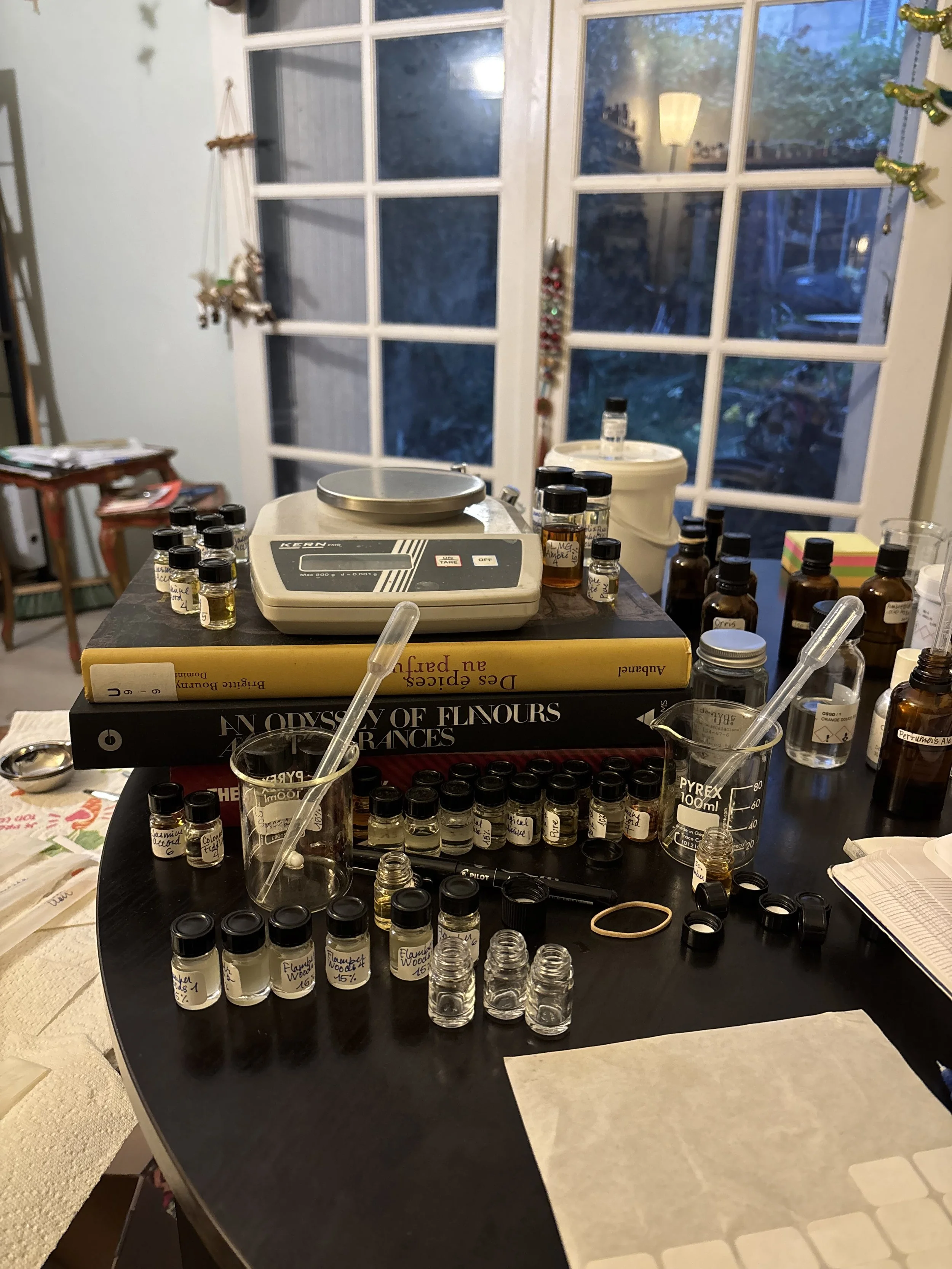 Table de travail avec plusieurs fioles de différentes tailles contenant des huiles ou des ingrédients liquides, des livres, une balance de laboratoire, des pipettes, un bécher gradué, un carnet de notes, et divers autres outils de laboratoire. En arrière-plan, des fenêtres laissent passer la lumière naturelle dans une pièce.