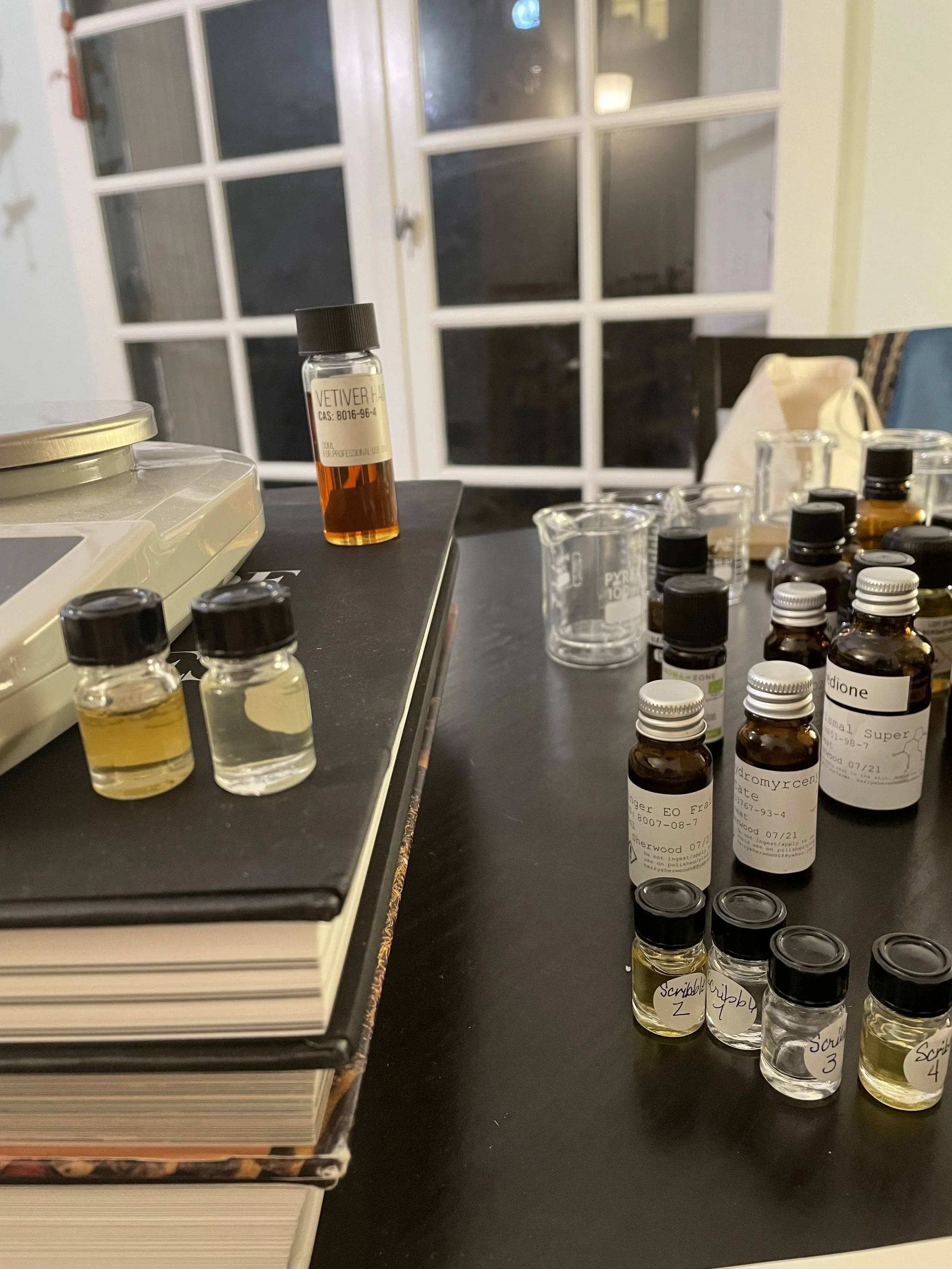 Une table avec plusieurs petites fioles contenant des liquides de différentes couleurs, une balance, des livres empilés, et des tasses en verre avec des outils de laboratoire. Fenêtre en arrière-plan.