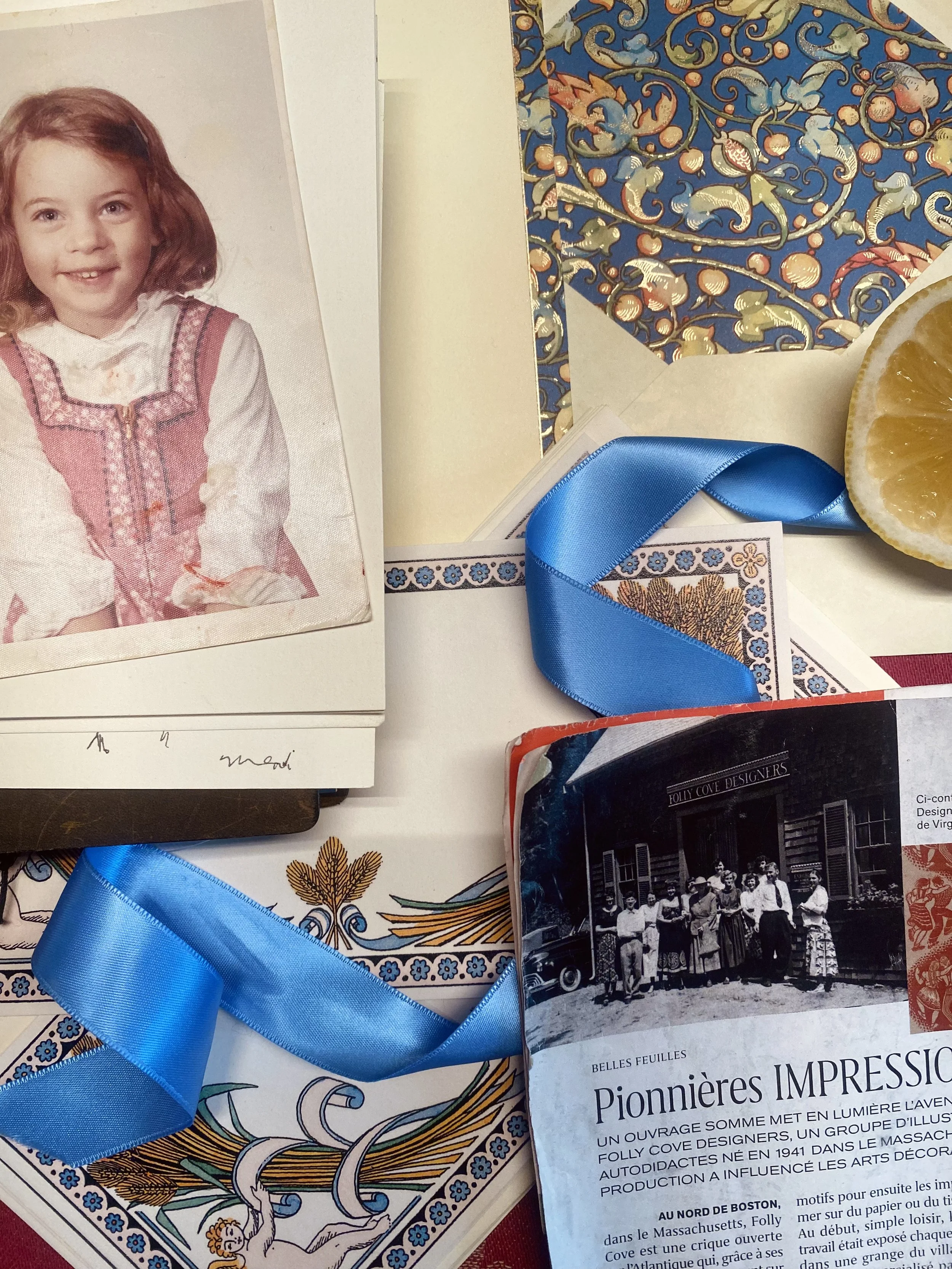 Vintage childhood photo, French book, blue ribbon, botanical paper, and citrus slice arranged in artistic flat lay about scent memory and perfumery.