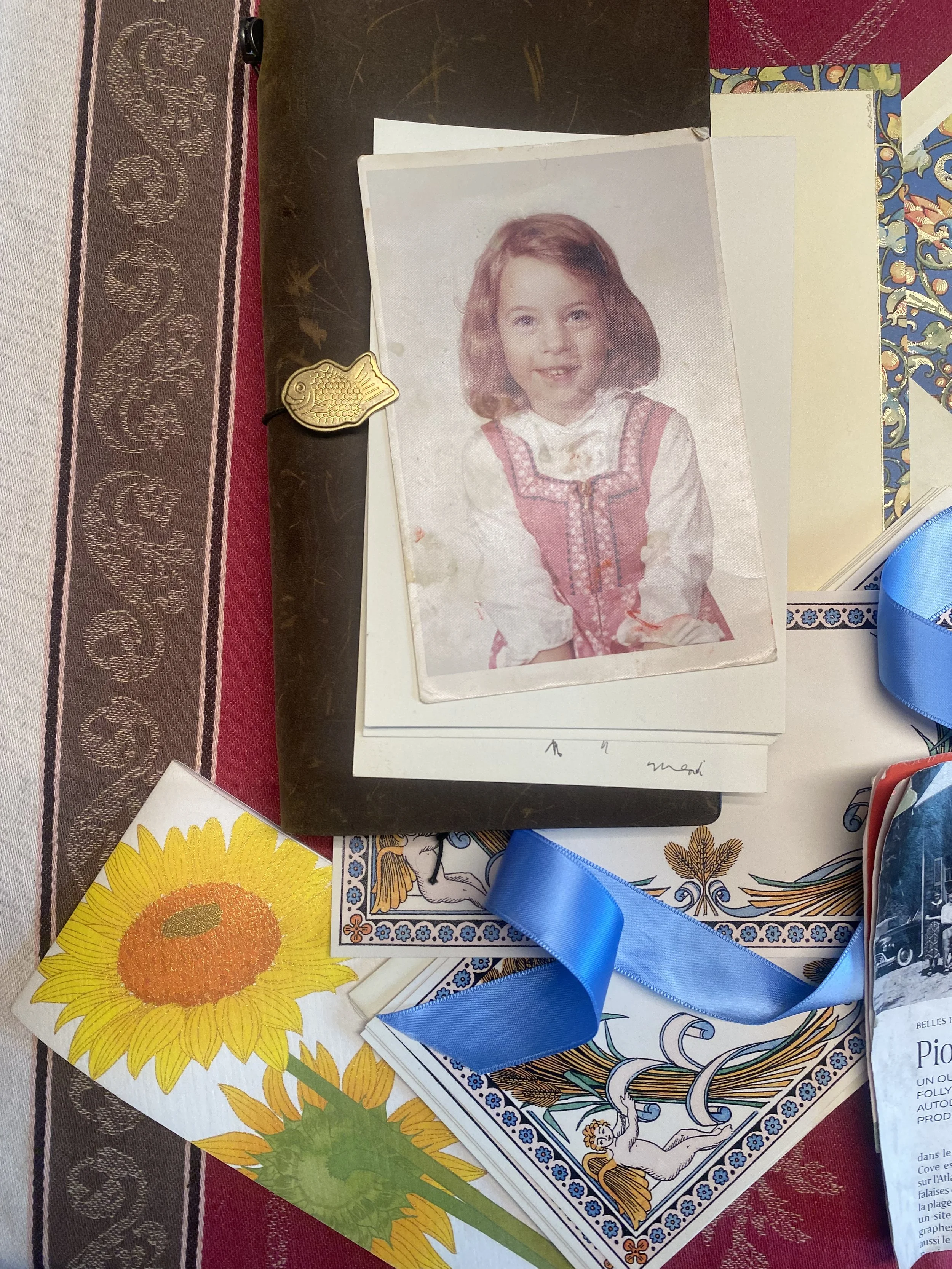 Une vieille photo d'une jeune fille avec des cheveux bruns, portant une robe traditionnellement russe, est posée sur une table avec des broderies, une carte de lecture, et un ruban bleu.