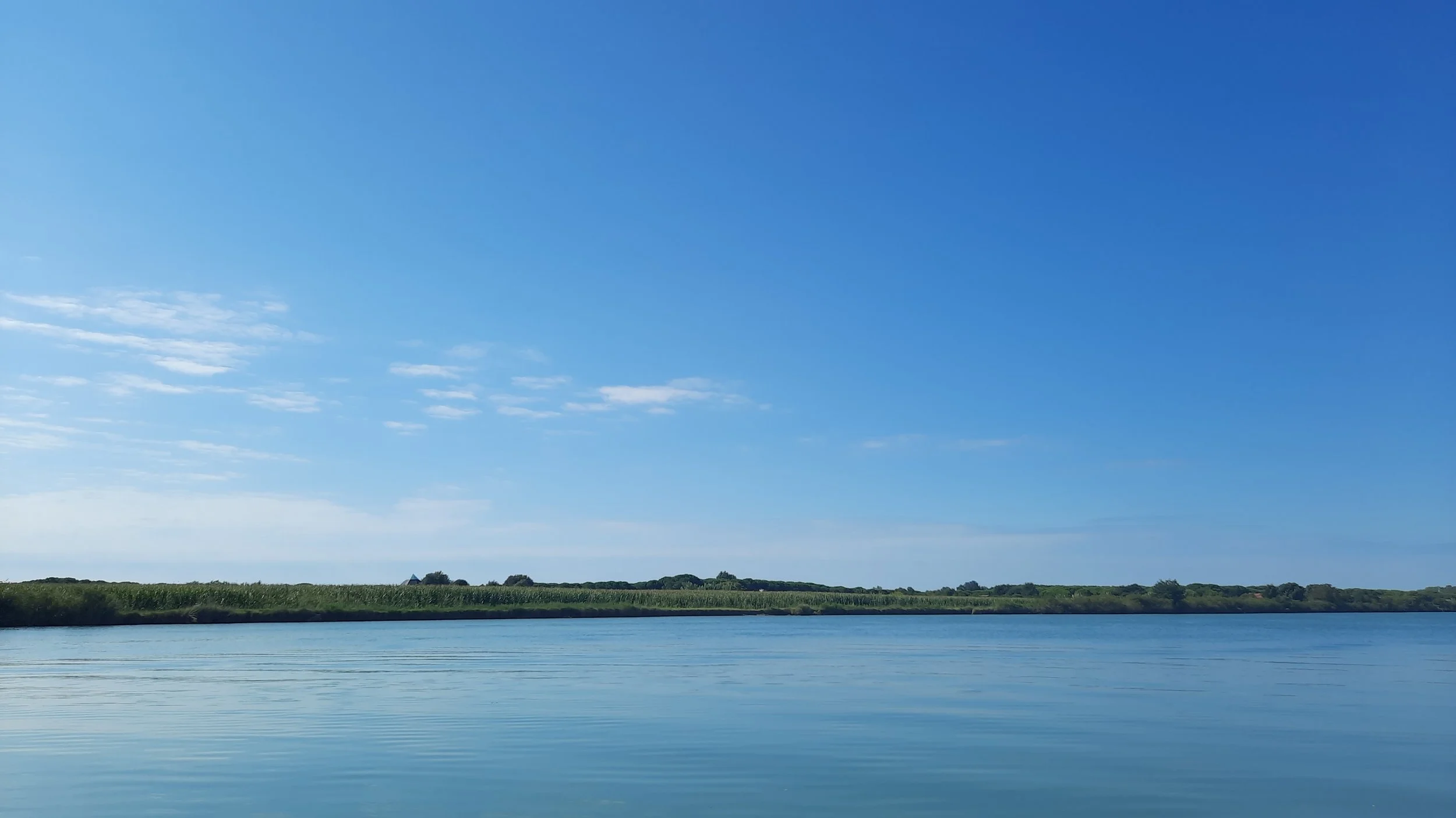 Paysage de rivière avec un ciel bleu clair et quelques nuages, avec des végétations verdoyantes le long de la rive.
