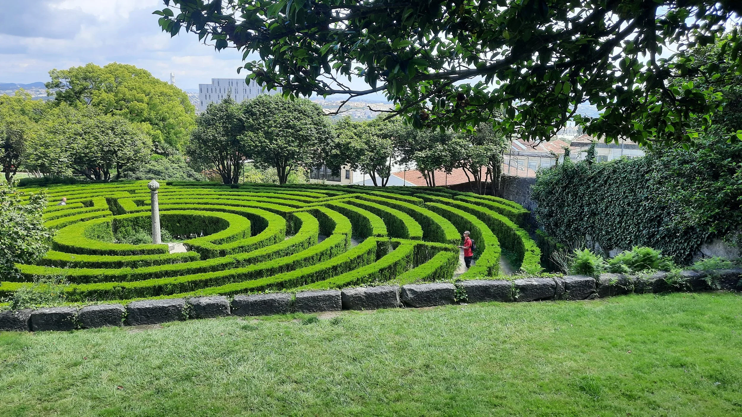 Jardin avec des haies en forme de labyrinthes, un homme portant un chapeau et un sac à dos. Arbres verts et bâtiments en ville en arrière-plan.