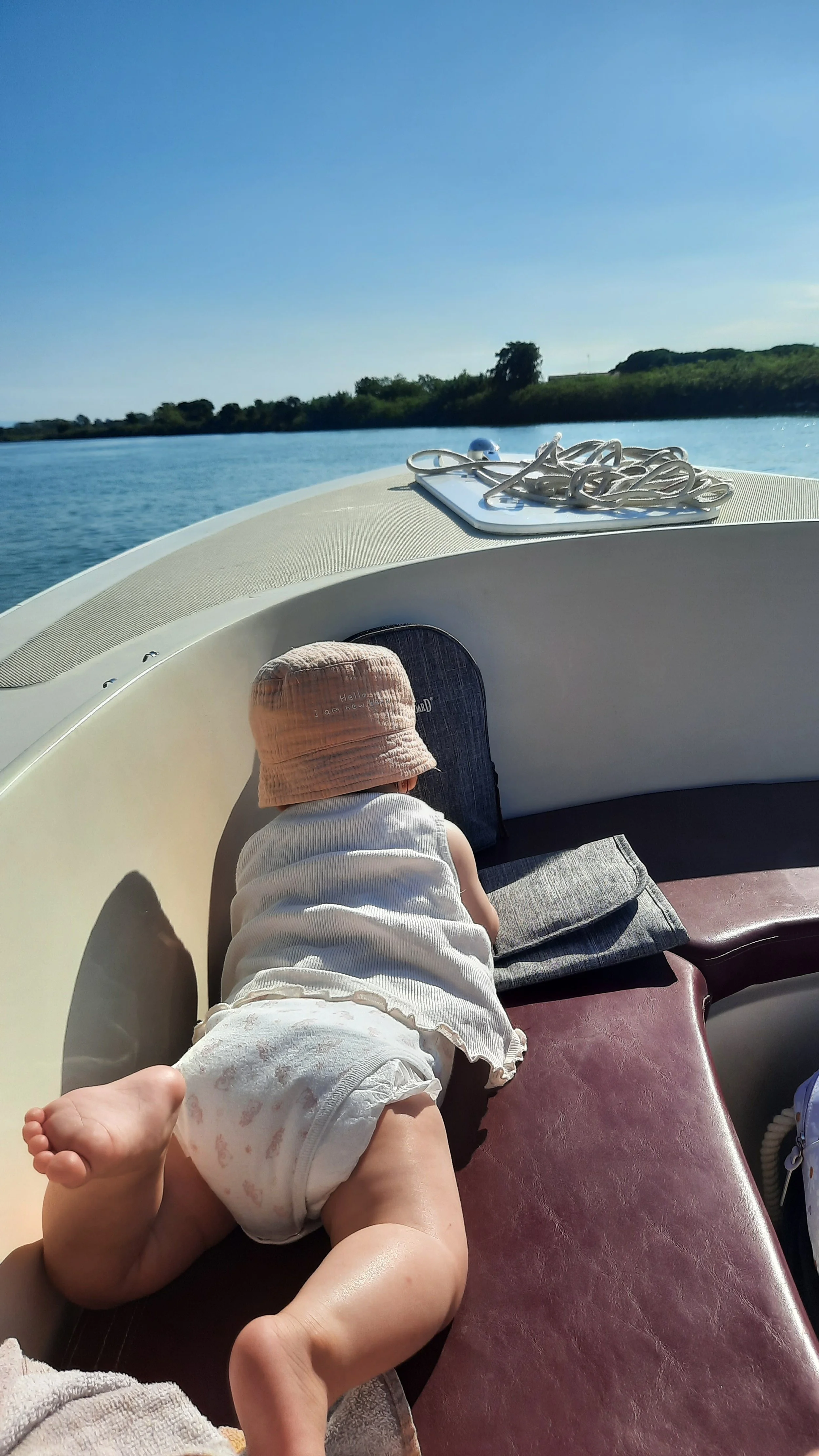 Un bébé en couche et chapeau blanc allongé à l'avant d'un bateau, avec un paysage de rivière et de nature en arrière-plan.