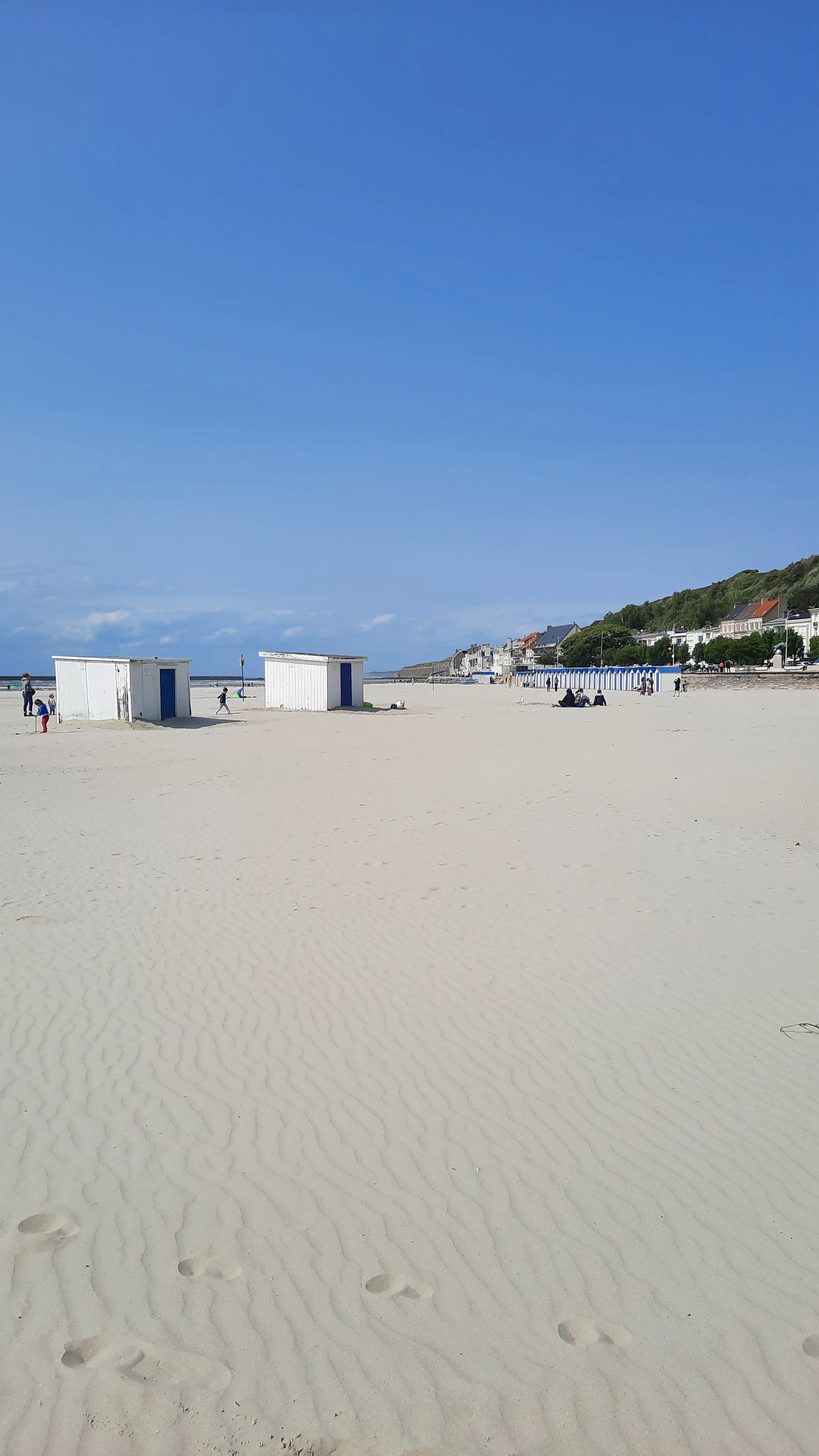 Plage de sable avec quelques cabanes blanches, des personnes au loin, un ciel bleu et des bâtiments en arrière-plan.