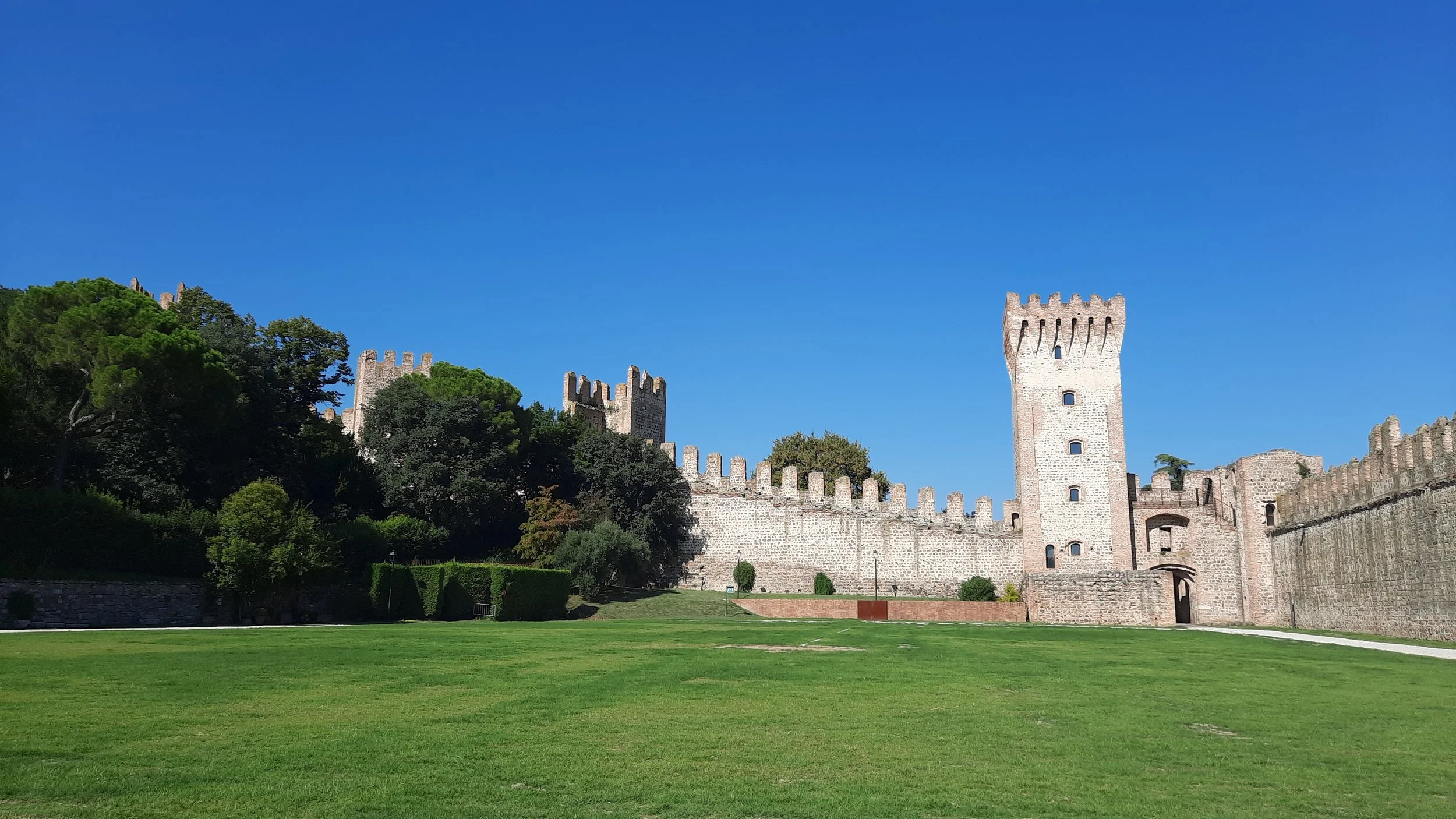 Muraille et tours d'un château ancien entouré d'un espace herbeux et d'arbres, sous un ciel clair et bleu.