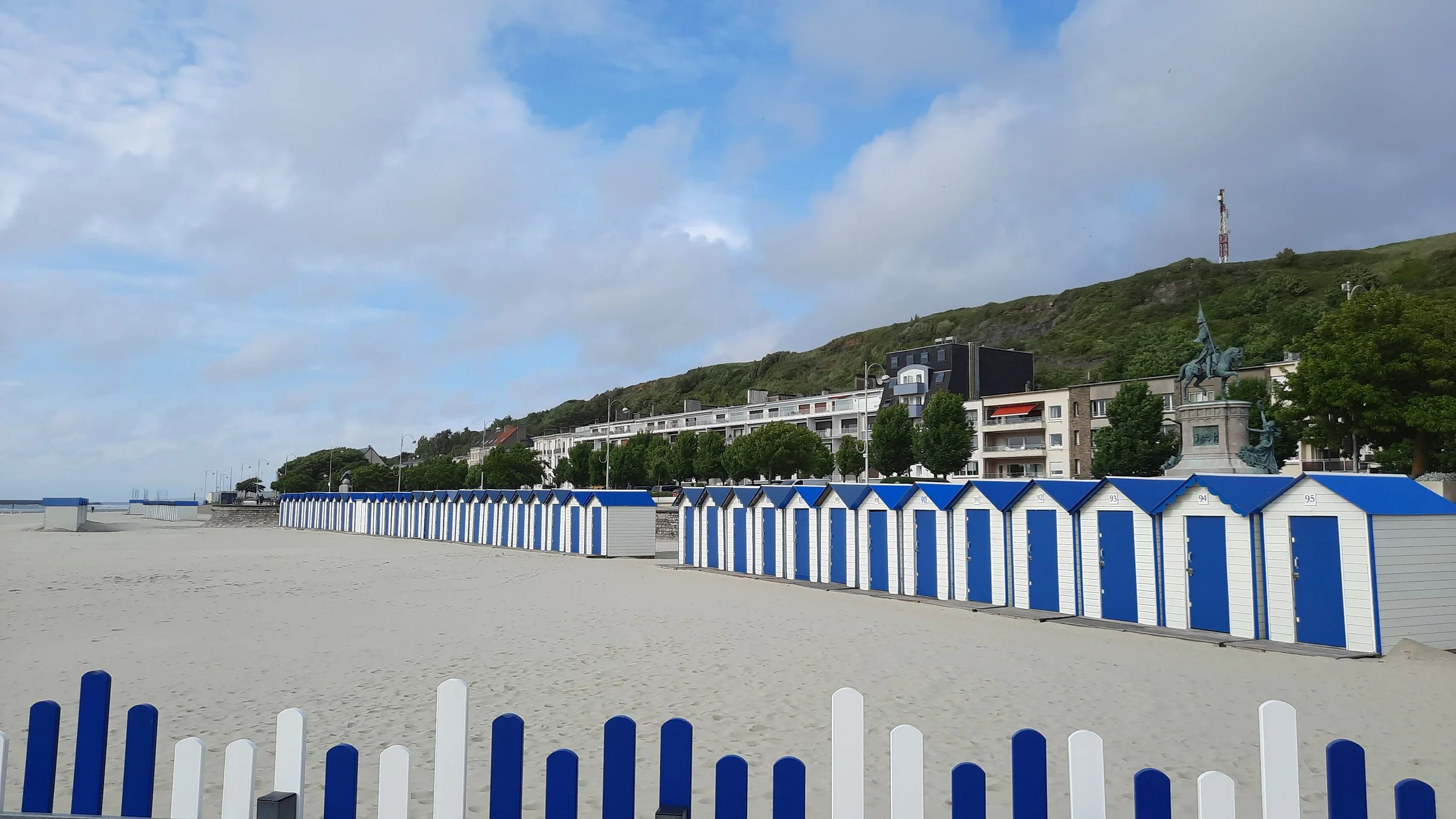 Plage avec cabines de plage bleues et blanches, statues, bâtiments résidentiels en arrière-plan, collines verdoyantes, ciel partiellement nuageux.