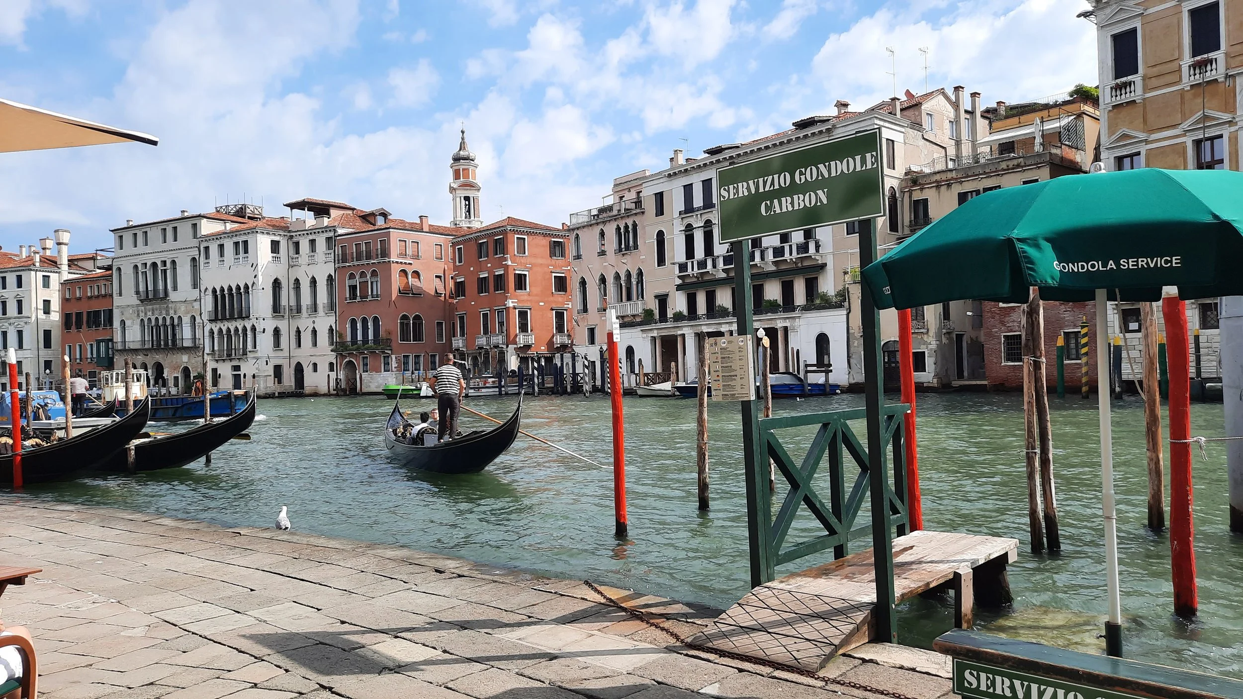 Canaux de Venise avec gondoles, bâtiments historiques colorés en arrière-plan, et un panneau indiquant un service de gondole en italien.