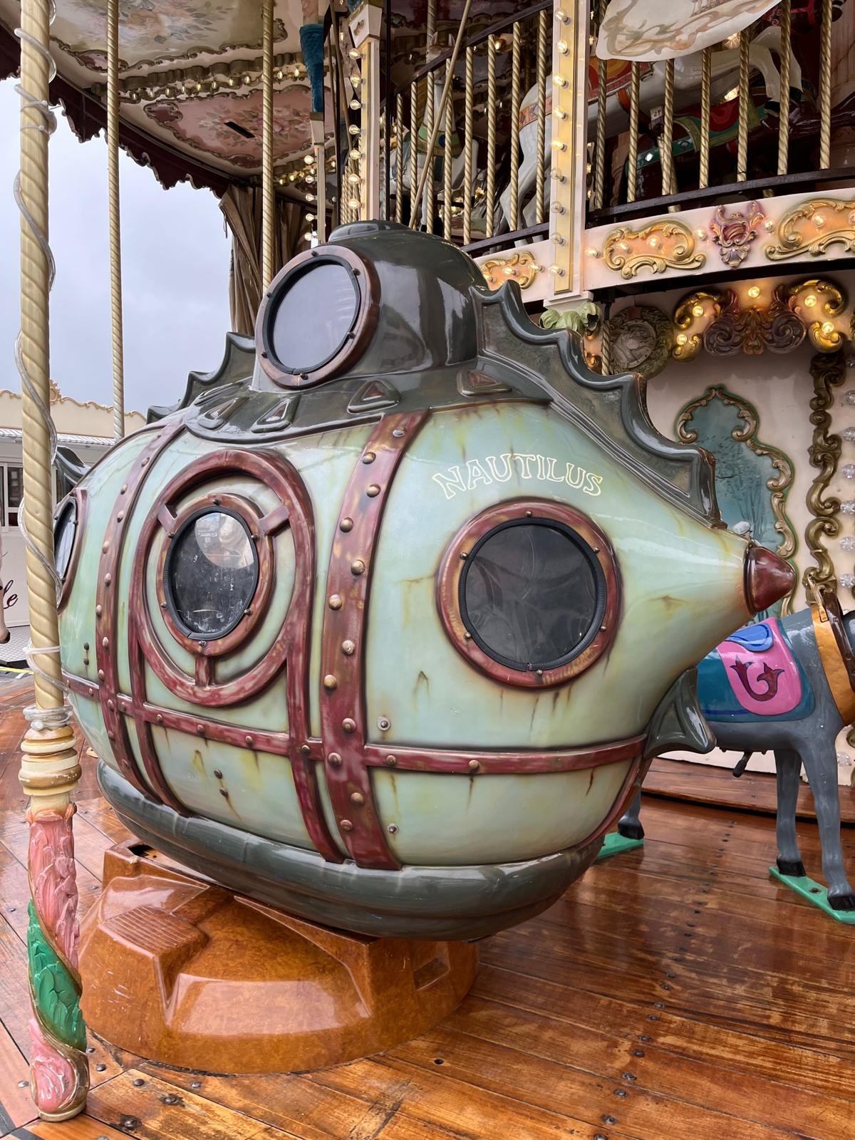 Une attraction de manège en forme de sous-marin vintage avec le nom 'Nautilus' écrit dessus, situé sur un parquet en bois, avec un deuxième étage de manège décoré en arrière-plan.