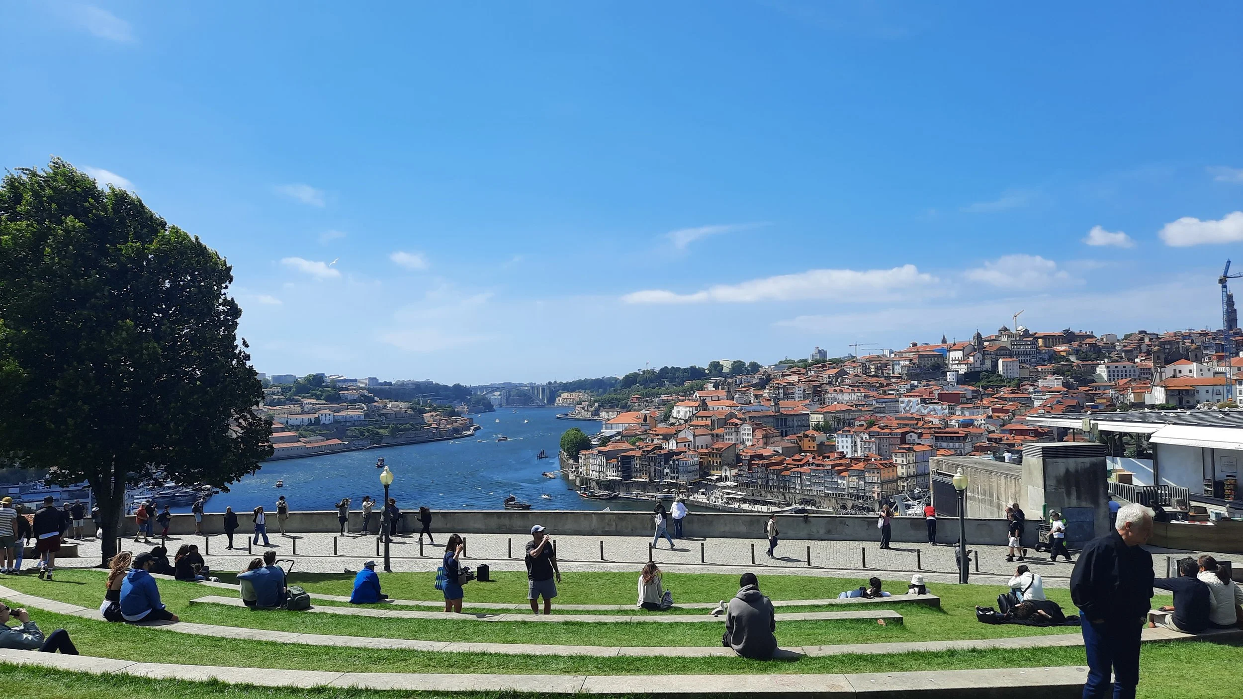 Une vue panoramique d'une ville construite sur une colline, avec un fleuve en contrebas, des ponts, et des bâtiments aux toits rouges sous un ciel bleu avec quelques nuages, vue depuis un parc où des personnes se détendent et se promènent.