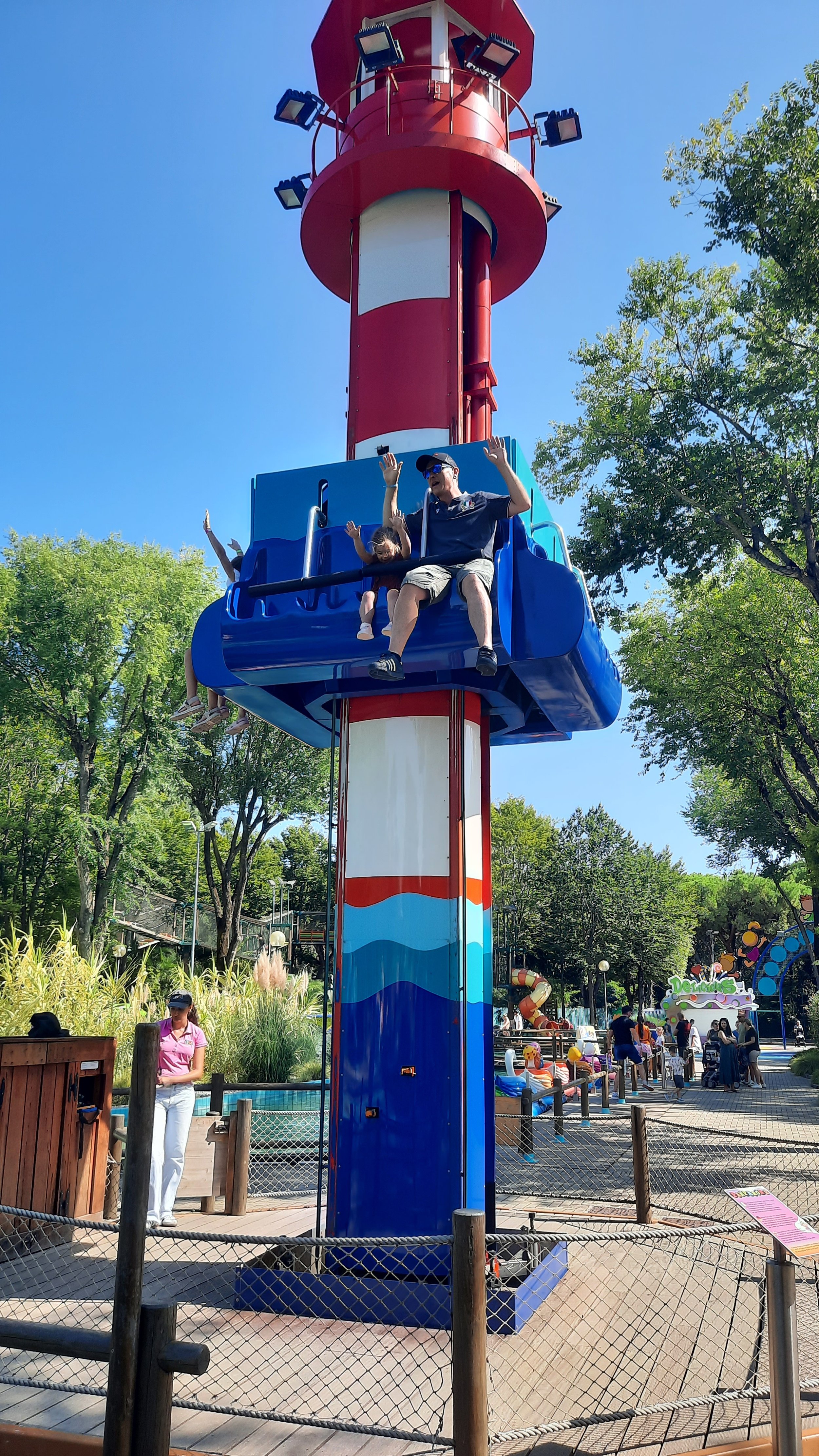 Des personnes s'amusent sur une tour de jeu en parc d'attractions, un homme et deux enfants souriants, avec des arbres et d'autres attractions visibles en arrière-plan.