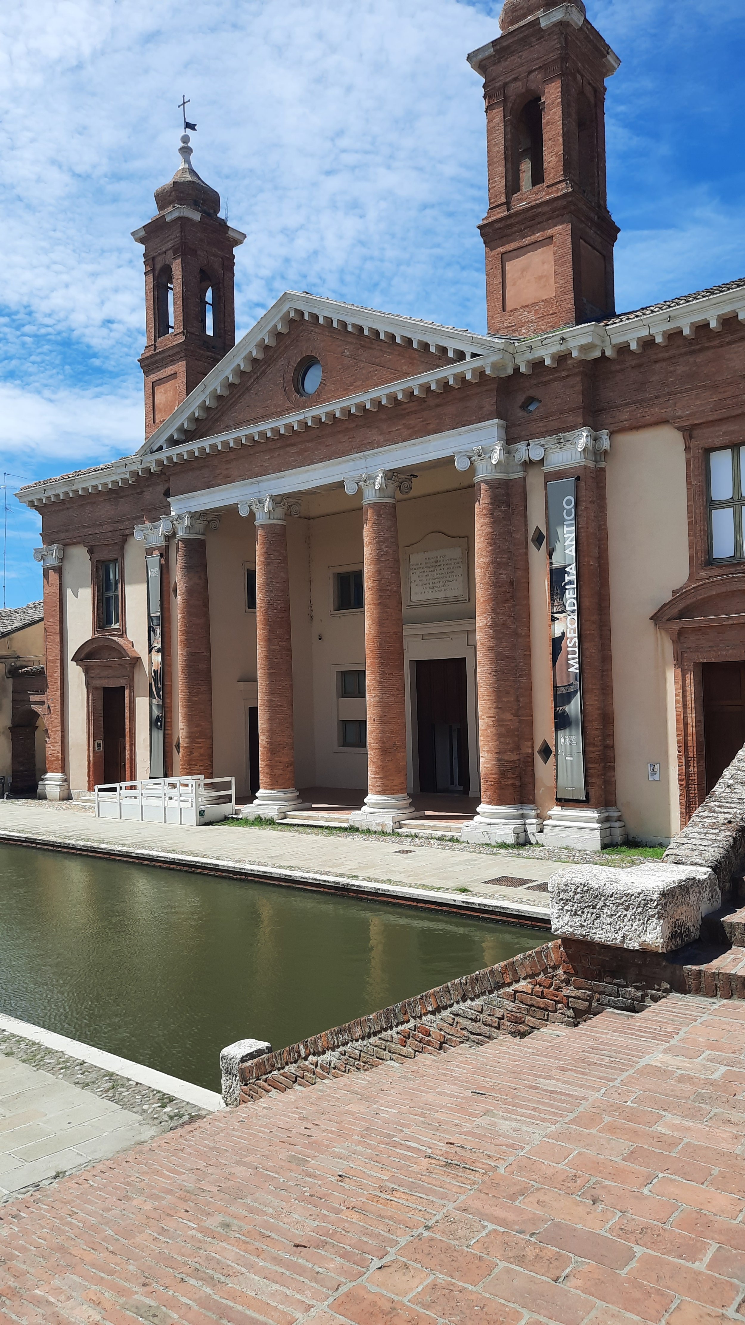 Une vieille bâtisse en briques avec deux clochers et d'amples colonnes colorées, située le long d'un canal.