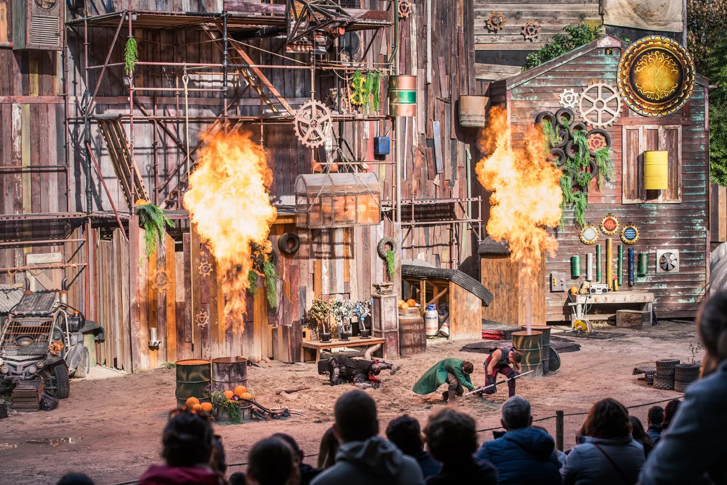 Spectacle de cascades avec flammes, personnes déguisées en pirates, décor rustique en bois, et public regardant l'événement.