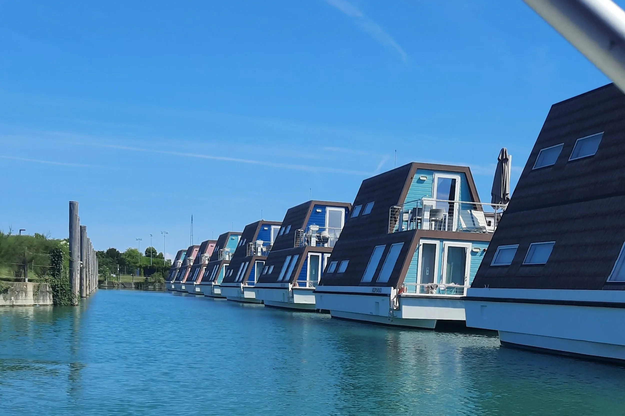 Une rangée de maisons flottantes colorées sur un canal, avec un ciel clair et du vert en arrière-plan.