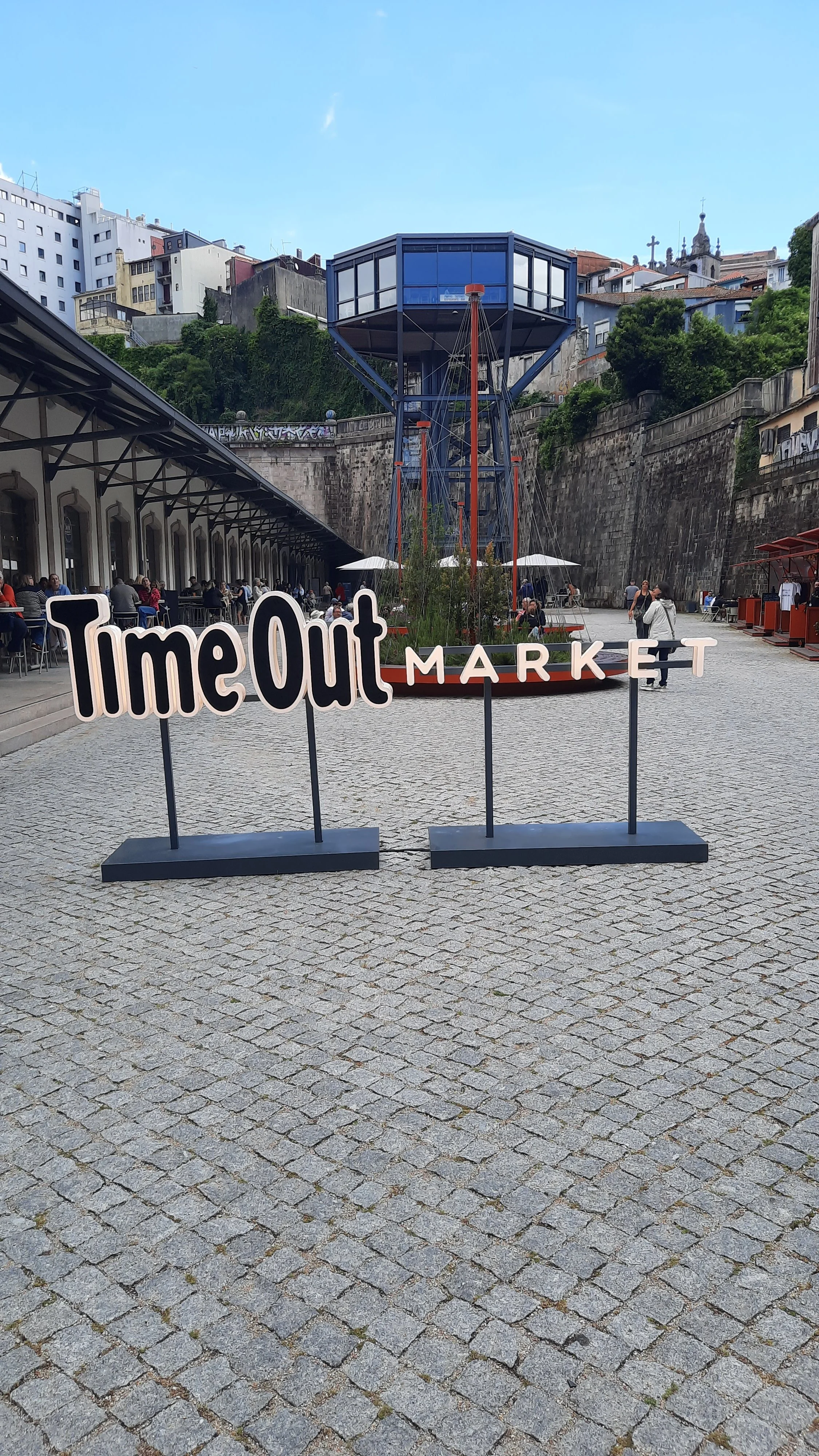 Entrée de marché avec un panneau indiquant 'Time Out MARKET'; au fond, un espace avec des tables et des parasols, et une structure en forme de tour bleue avec fenestrations, située au sommet d'une plateforme en hauteur. La place est pavée de pavés en pierre et il y a des bâtiments en arrière-plan sur une colline.