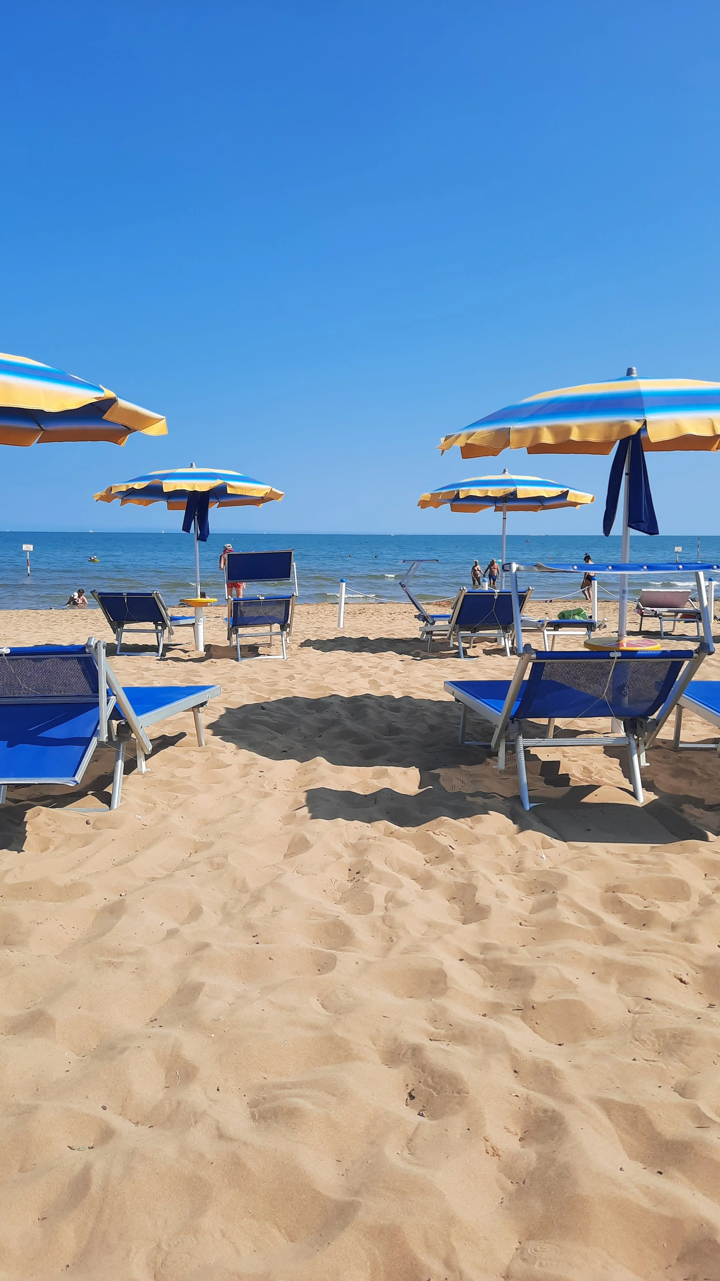 Plage ensoleillée avec des parasols jaunes et bleus, des chaises bleues, et des gens nageant ou se relaxant près de la mer.