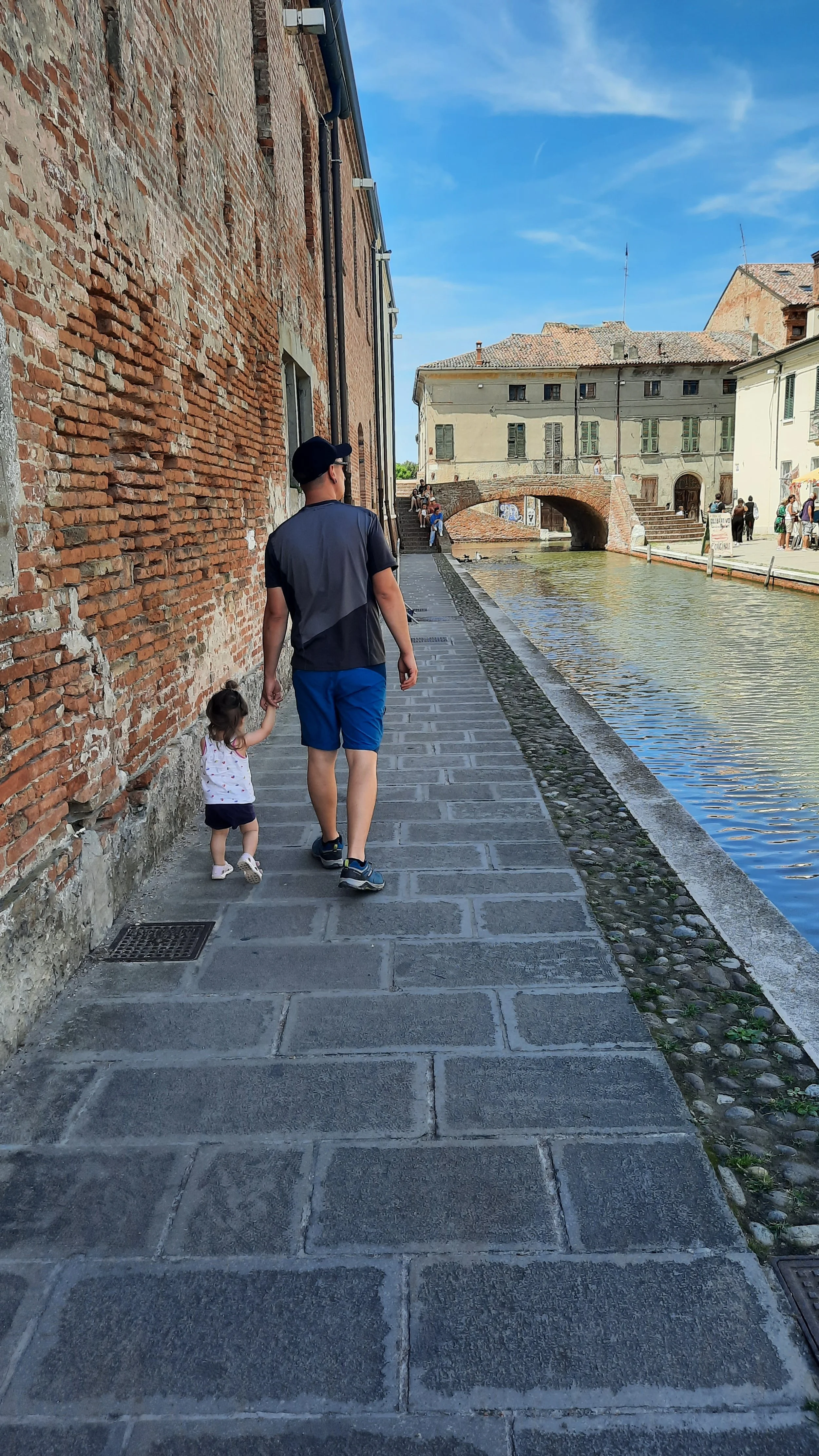 Un homme marche avec une jeune fille le long d'une voie d'eau bordée de bâtiments anciens en brique, sous un ciel bleu.