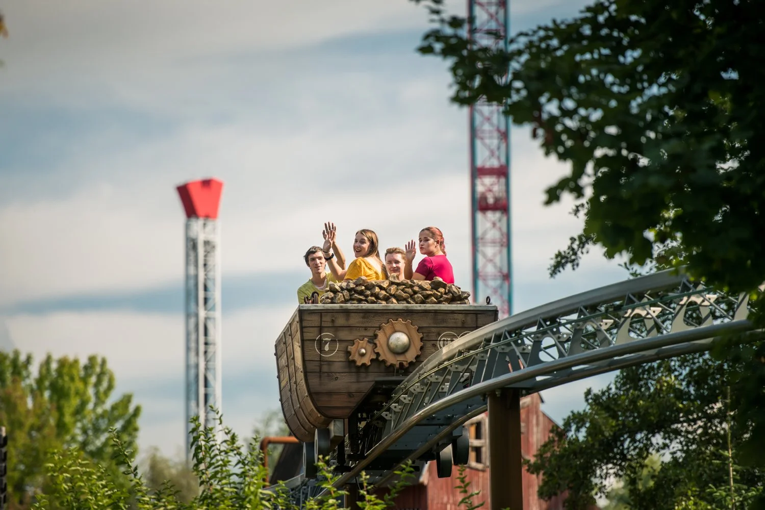 Groupe de quatre personnes dans une montagne russe en forme de bateau en bois, avec des engrenages décoratifs, passant devant des structures de montagnes russes en métal et entourées de verdure, sous un ciel nuageux.