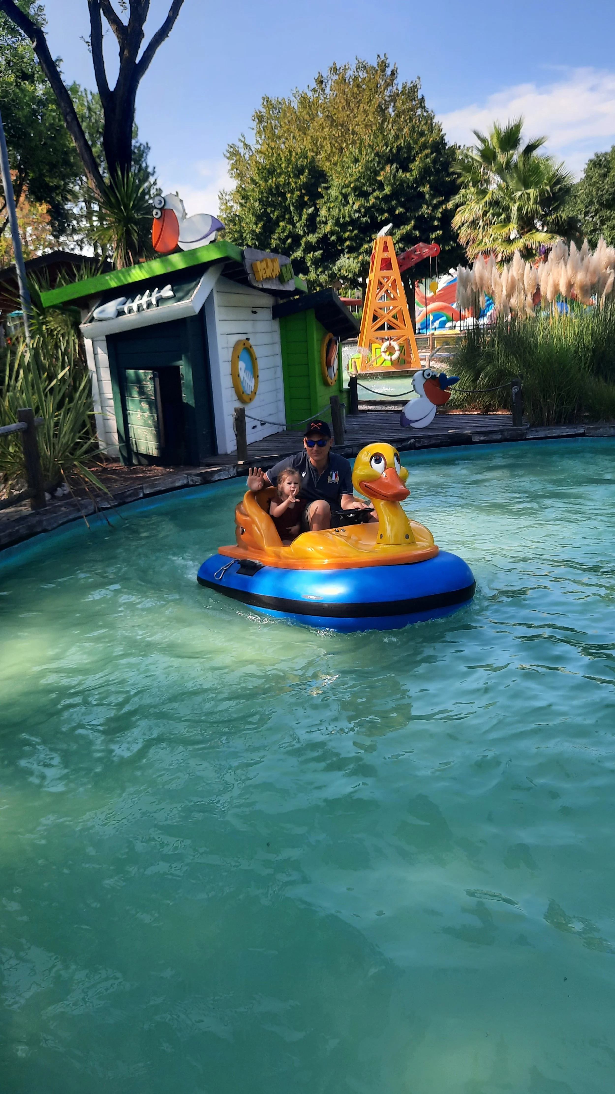 Une personne et un enfant dans une barque en forme de canard sur une attraction de parc d'attractions, avec un décor de mer et des éléments de plage en arrière-plan.