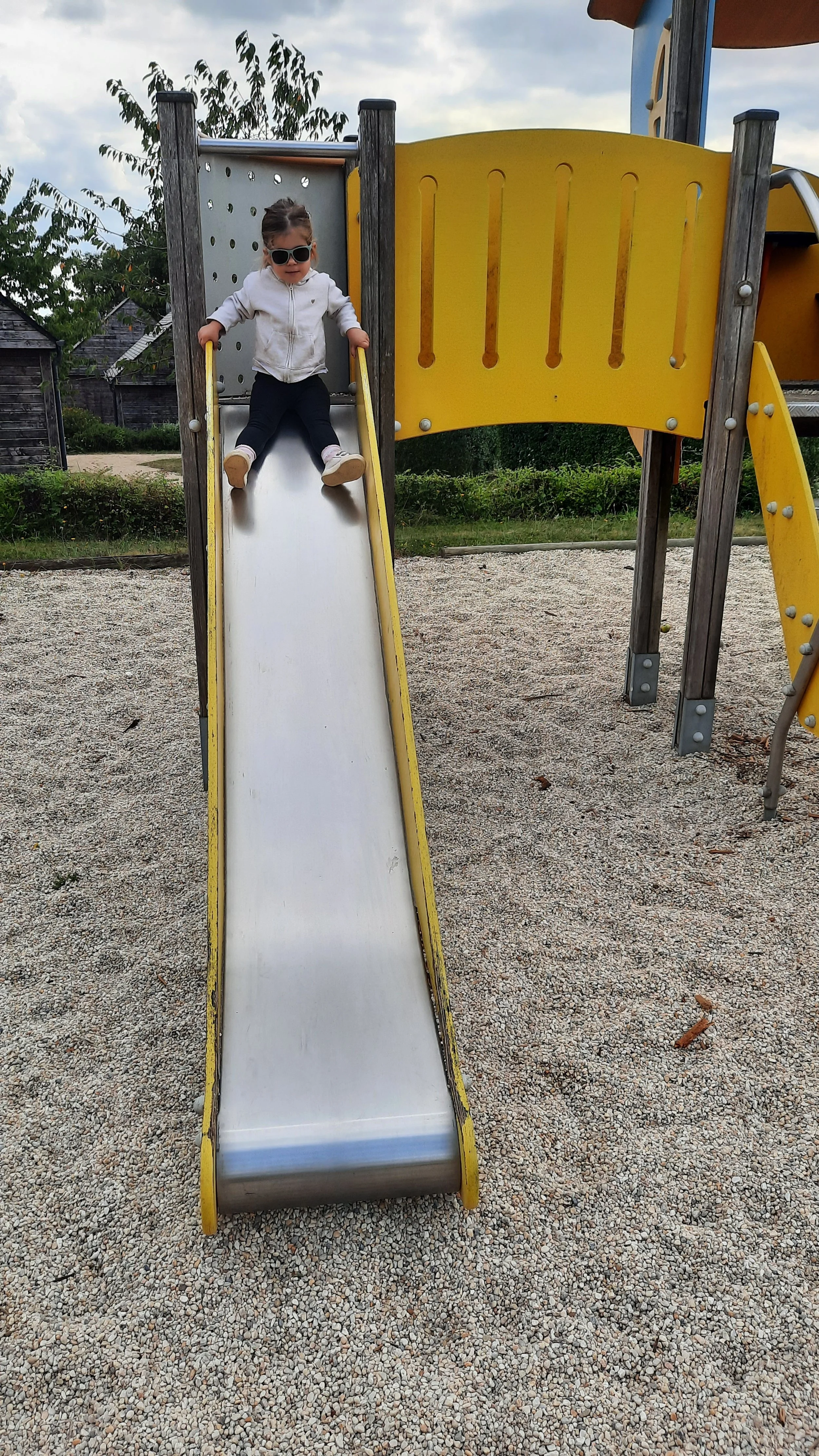 Une petite fille avec des lunettes de soleil descend ou monte une glissière dans une aire de jeux extérieure, avec des arbres et des bâtiments en bois en arrière-plan.