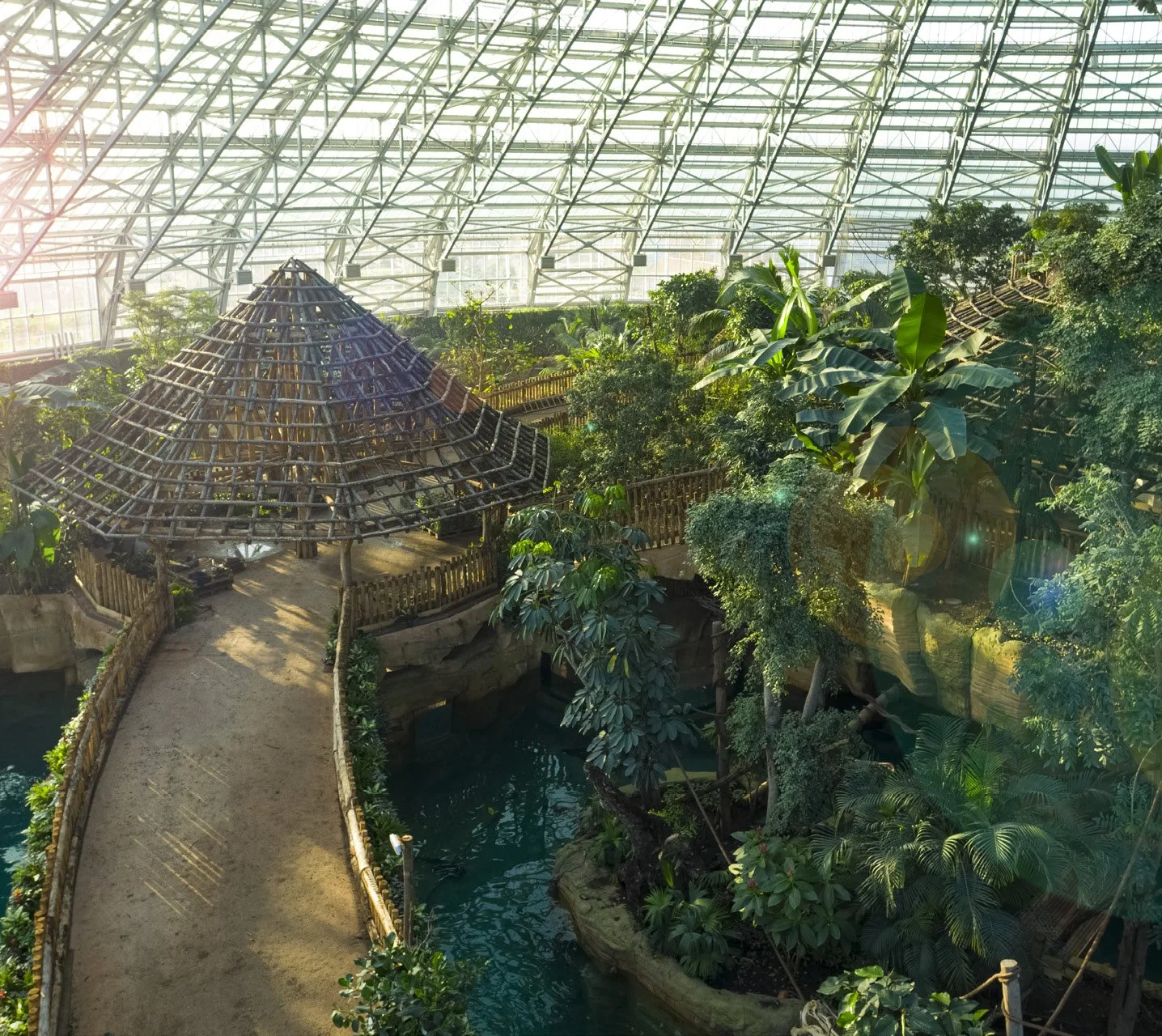 Intérieur d'une serre avec biodiversité, un chemin en bois, un gazebo en bois, une petite rivière, et de nombreuses plantes tropicales, sous une structure en verre.