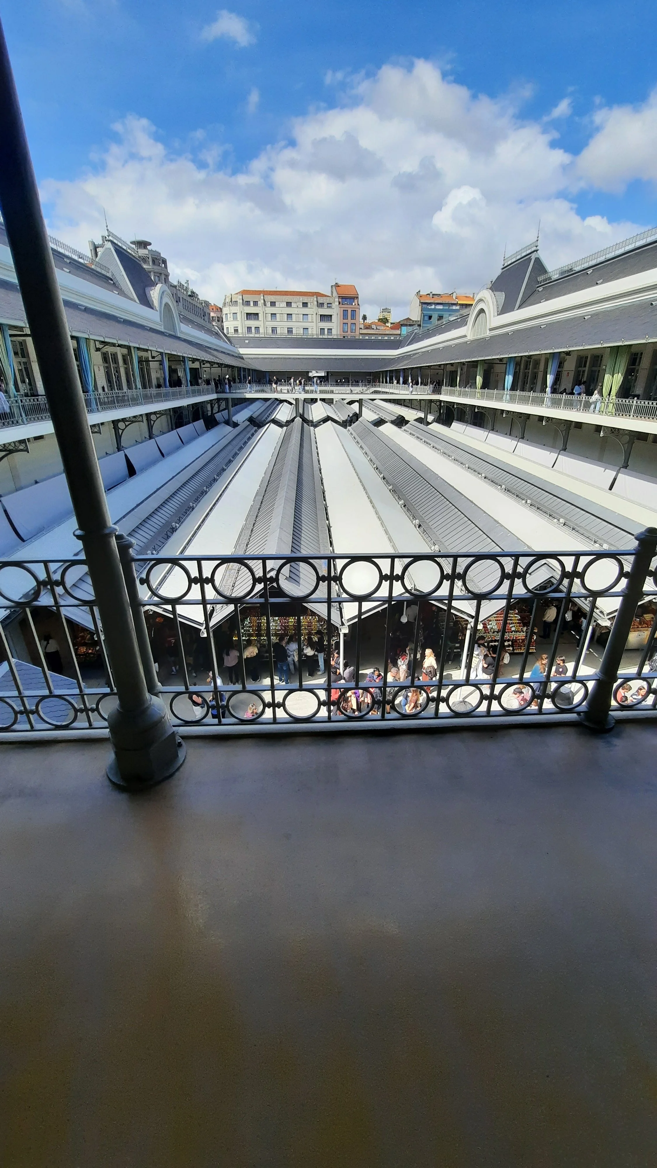 Vue depuis une fenêtre d'un bâtiment montrant une cour intérieure avec des étals de marché, des passants et des bâtiments en arrière-plan, sous un ciel partiellement nuageux.
