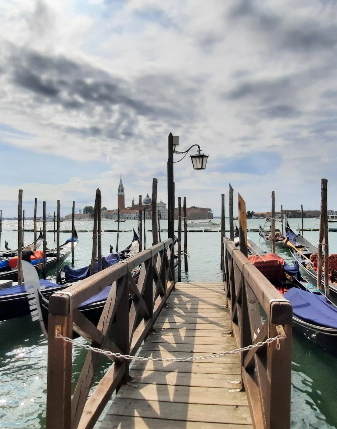 Quai en bois avec gondoles amarrées, vue vers la ville de Venise avec campanile et bâtiments historiques, ciel nuageux.