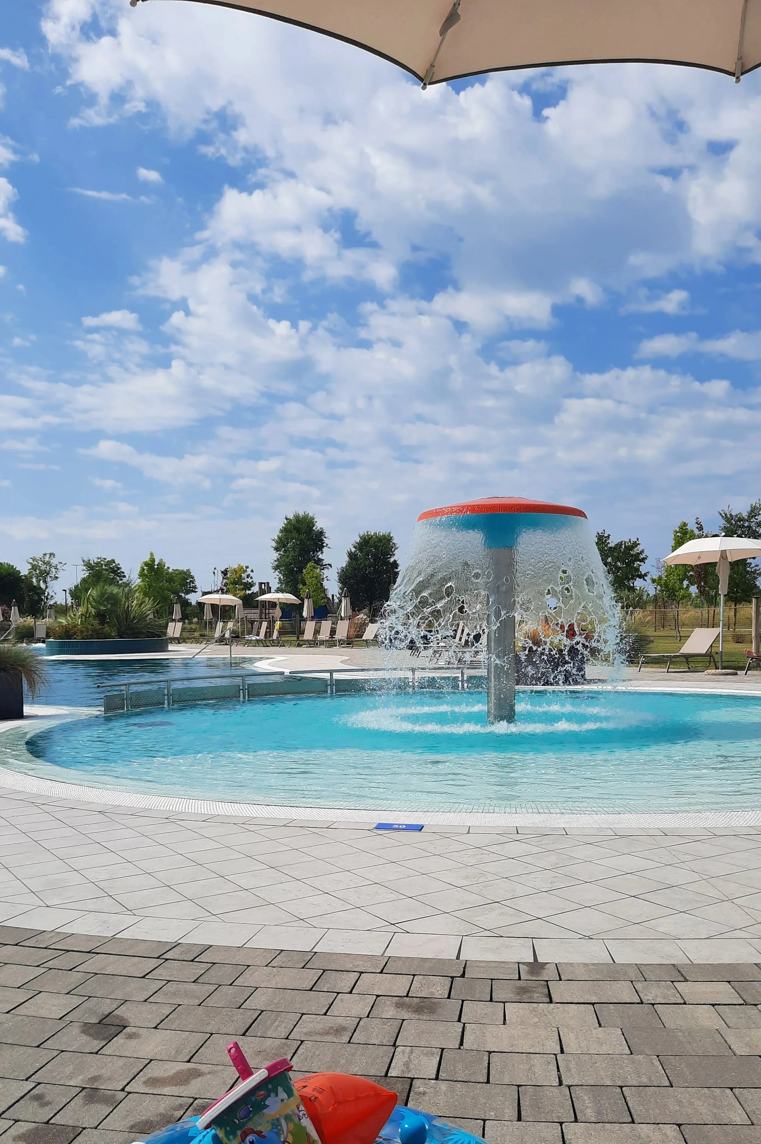 Piscine extérieure avec une fontaine d'eau en forme de champignon, parasols et chaises longues sous un ciel partiellement nuageux.