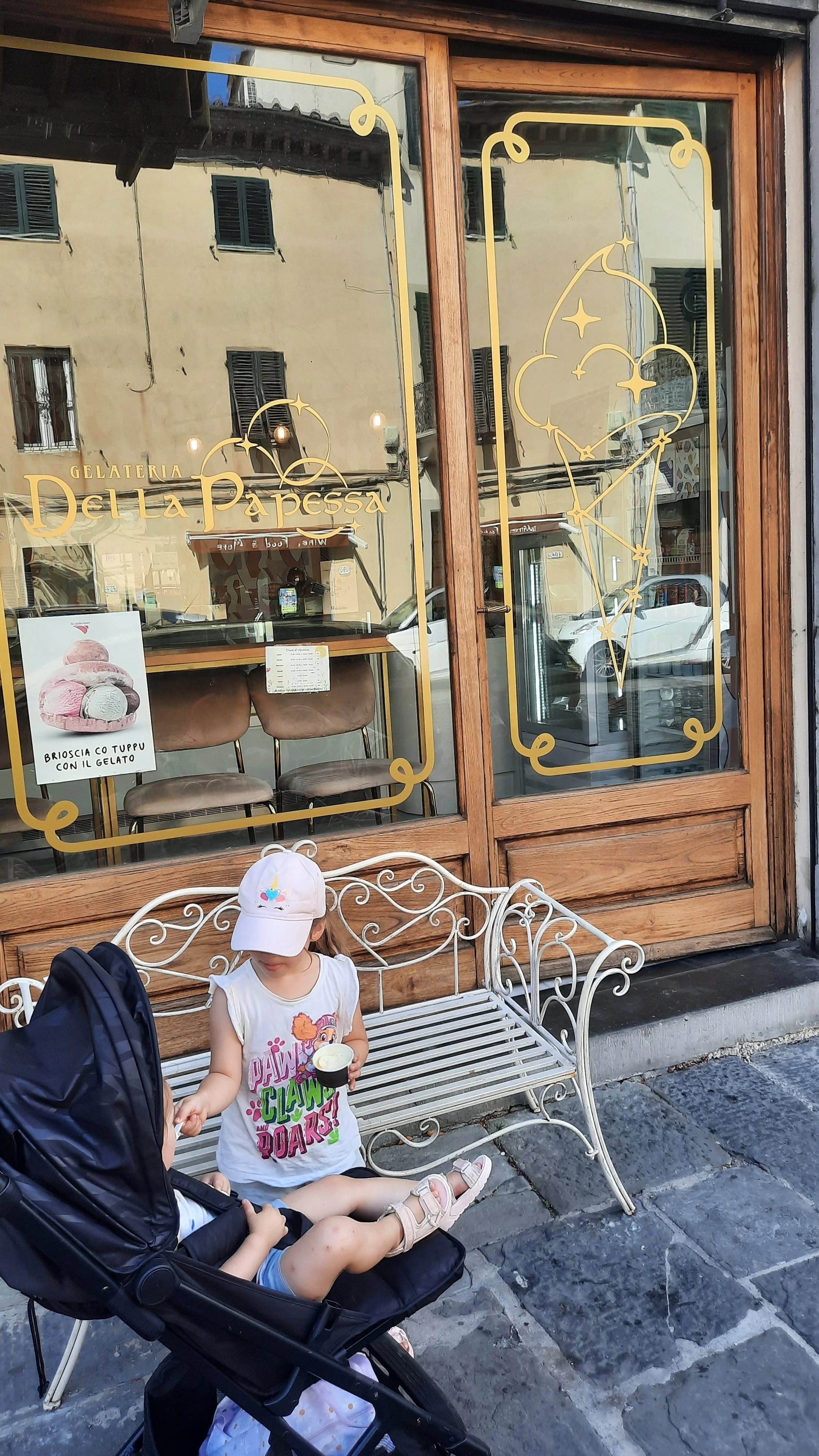 Une façade de gelateria avec des fenêtres en bois, décorée avec des illustrations de croissants de lune et d'étoiles, et un panneau promouvant des glaces à la noisette et au chocolat. Deux enfants sont assis sur un banc en métal blanc devant la boutique, une d'elles est dans une poussette, portant un t-shirt avec des dinosaures et une casquette blanche, et tenant une petite glace.