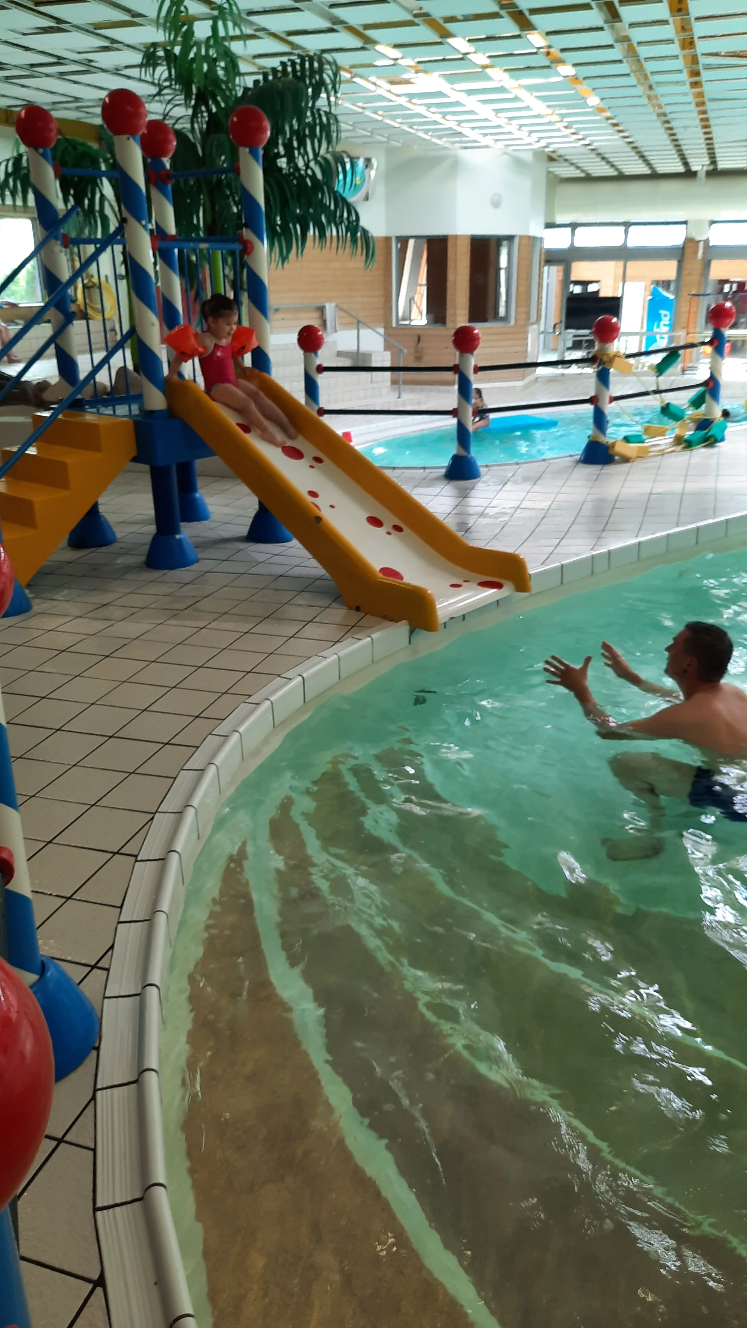 Une piscine intérieure avec un toboggan pour enfants, où une jeune fille est assise en haut et un homme nage dans l'eau à proximité.