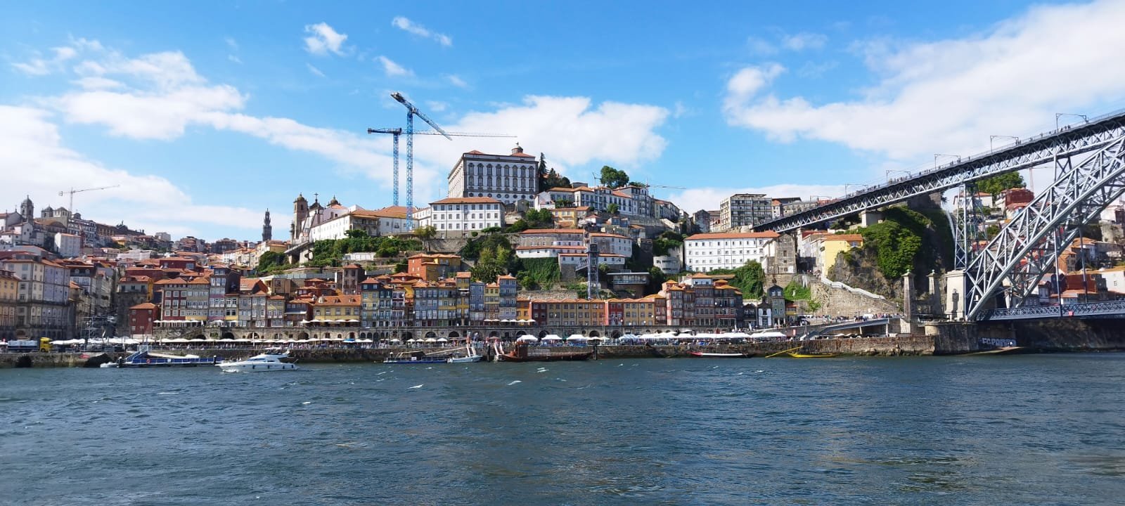 Vue d'une ville côtière avec des bâtiments colorés construits sur une colline, un pont en acier à droite et des bateaux dans l'eau au premier plan, sous un ciel bleu avec quelques nuages.