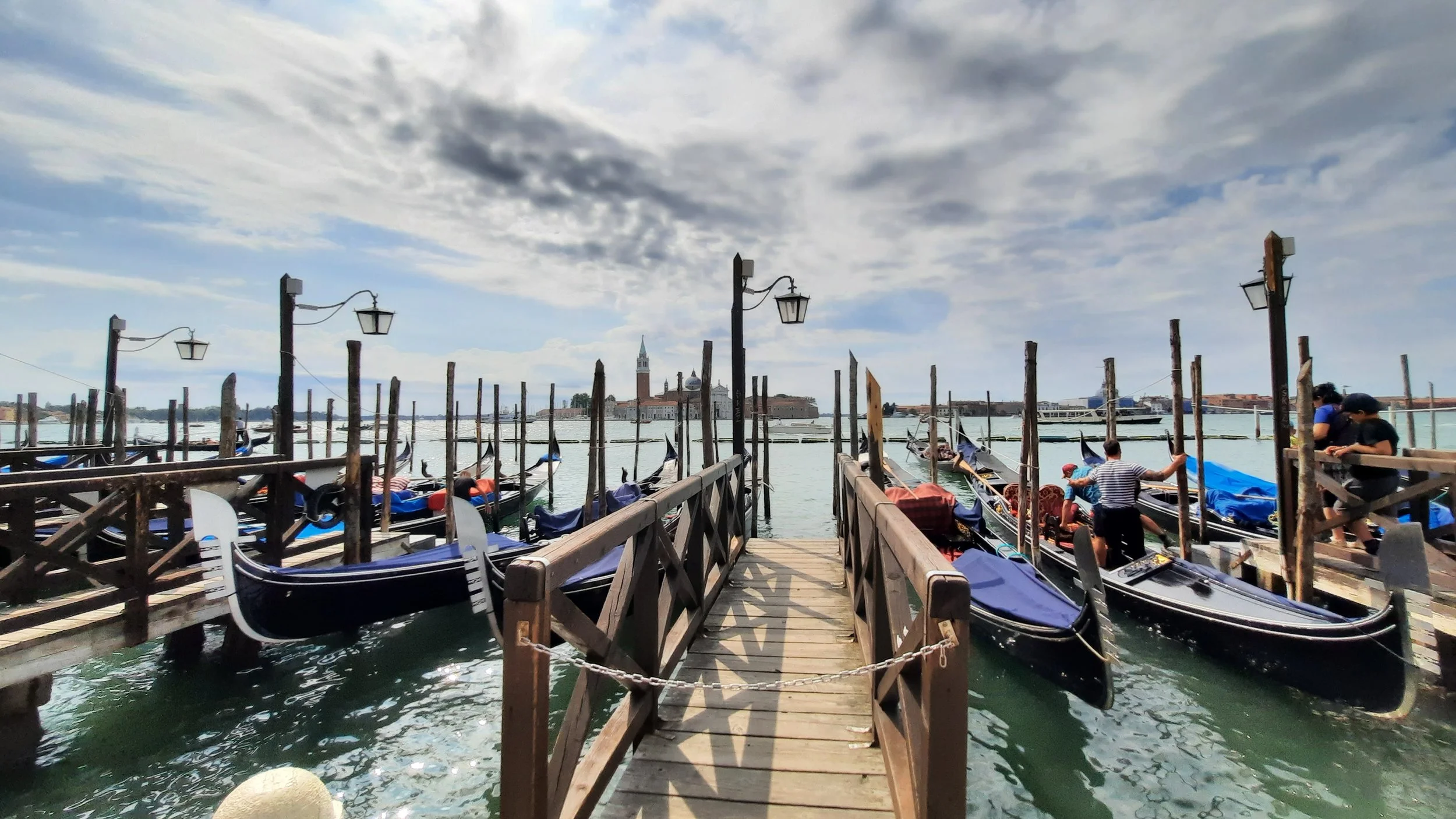 Petite jetée en bois menant à des quais avec des bateaux gondoles stationnés, fond avec bâtiments et cloché dans un ciel nuageux.