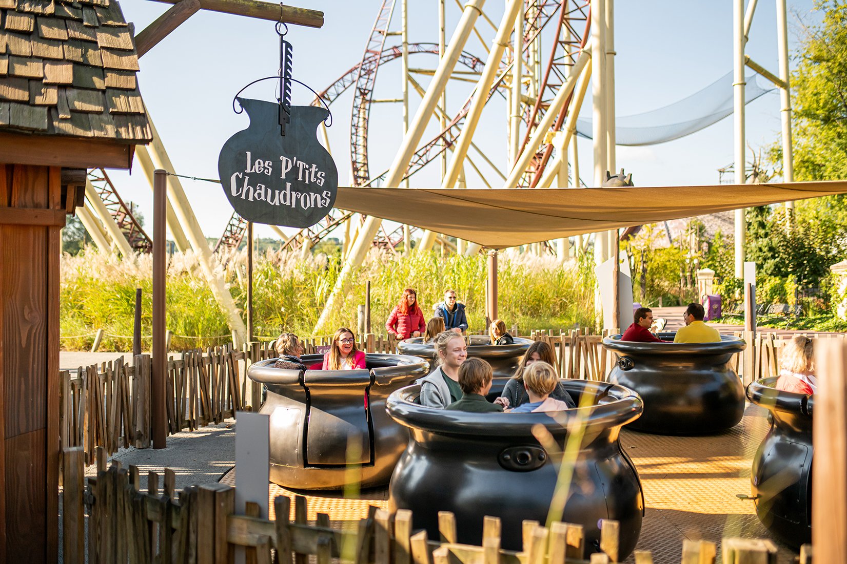 Des personnes en train de faire un tour en tasses de thé noir dans un parc d'attractions, avec des montagnes russes en arrière-plan et un panneau indiquant 'Les P'tits Chaudrons'.