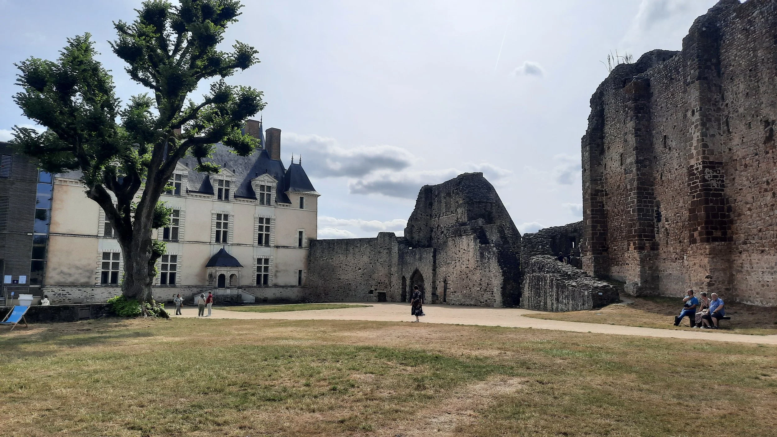 Une scène dans un parc avec un grand arbre, un bâtiment historique et des ruines en pierre, quelques personnes assises sur un banc, d'autres se promenant sous un ciel nuageux.