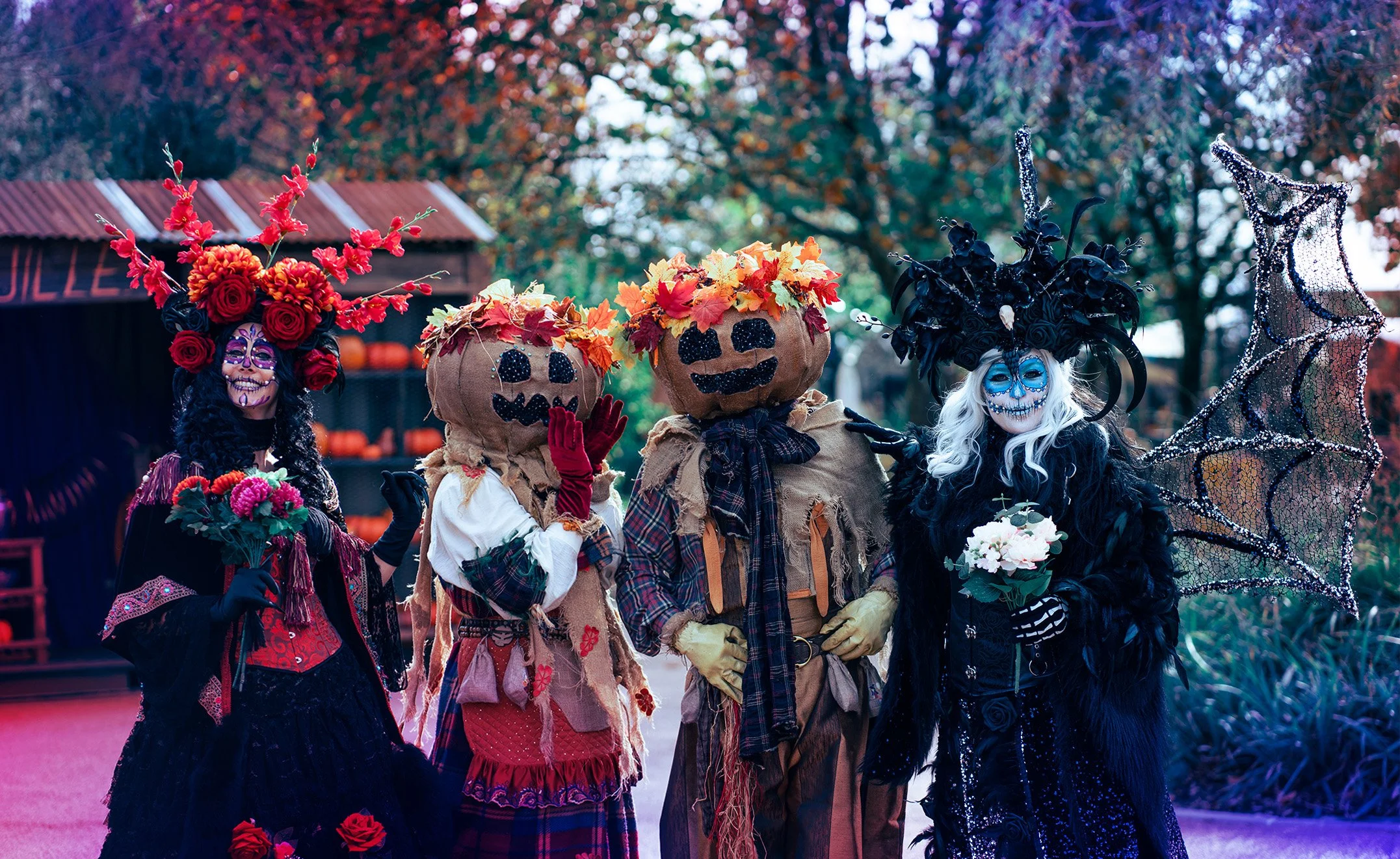 Personnages en costumes de Dia de los Muertos avec maquillages faciaux colorés, costumes élaborés, et accessoires thématiques, célébrant Halloween ou le Día de los Muertos dans un décor extérieur avec arbres et stand.