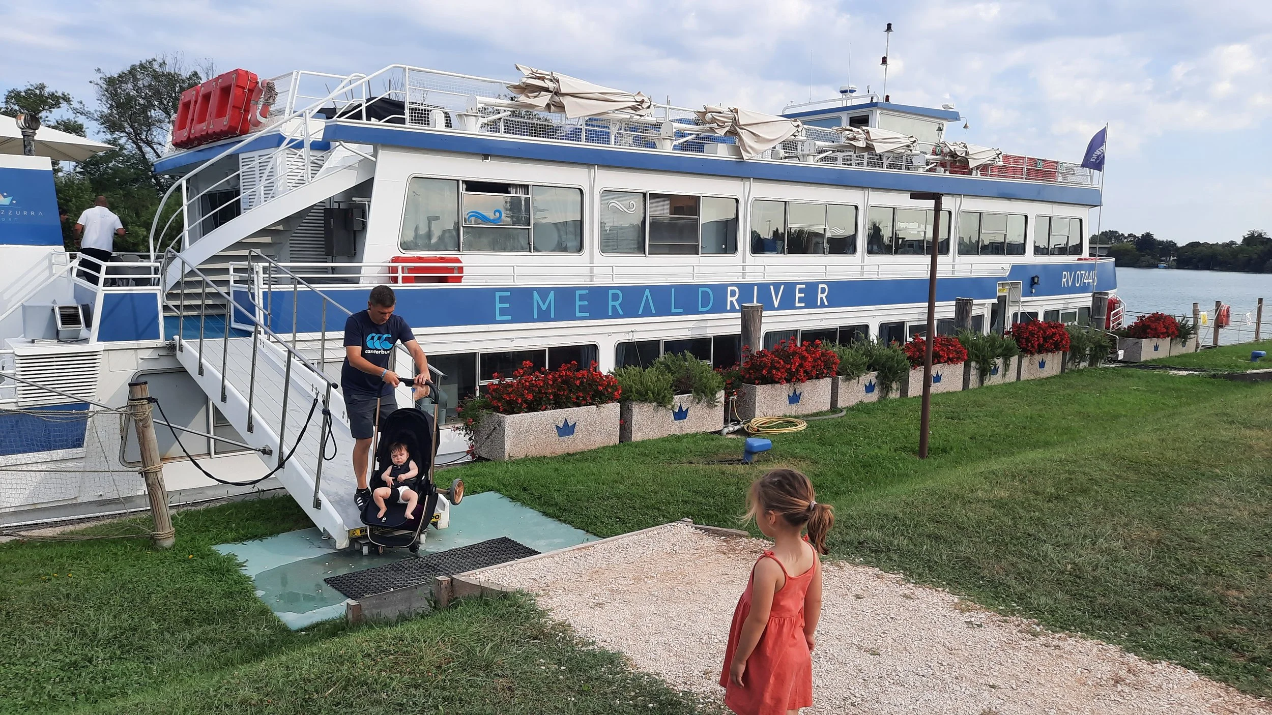 Un ferry nommé Emerald River est prêt à partir sur la rivière, avec des passagers, un homme poussant une poussette avec un jeune enfant, et une fille portant une robe orange se tenant près de l'eau, à côté de fleurs en pot