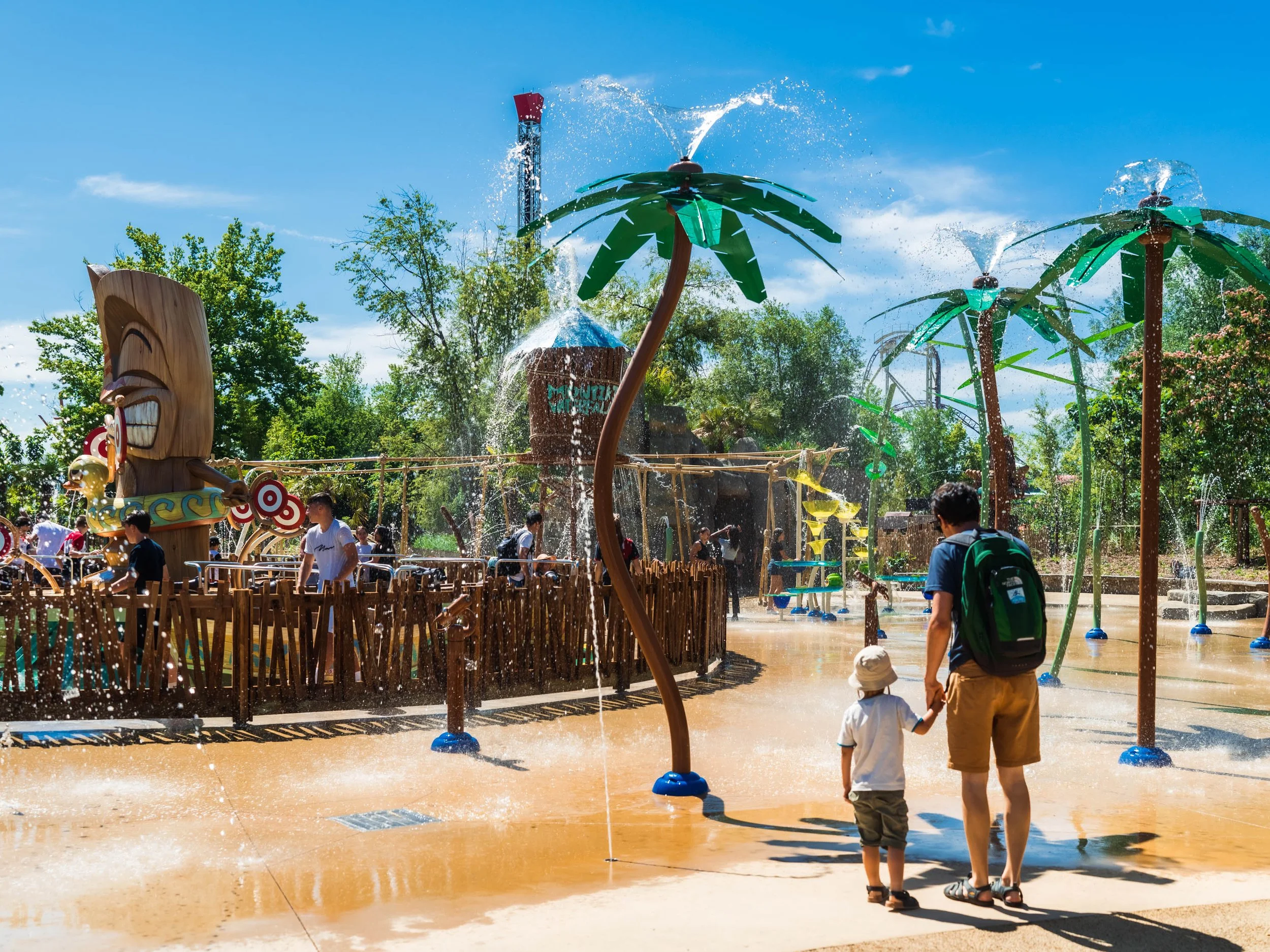 Enfants et adultes jouent dans une aire de jeux aquatique avec des fontaines, des palmiers en plastique, un totem en bois et des structures d'eau sous un ciel ensoleillé.